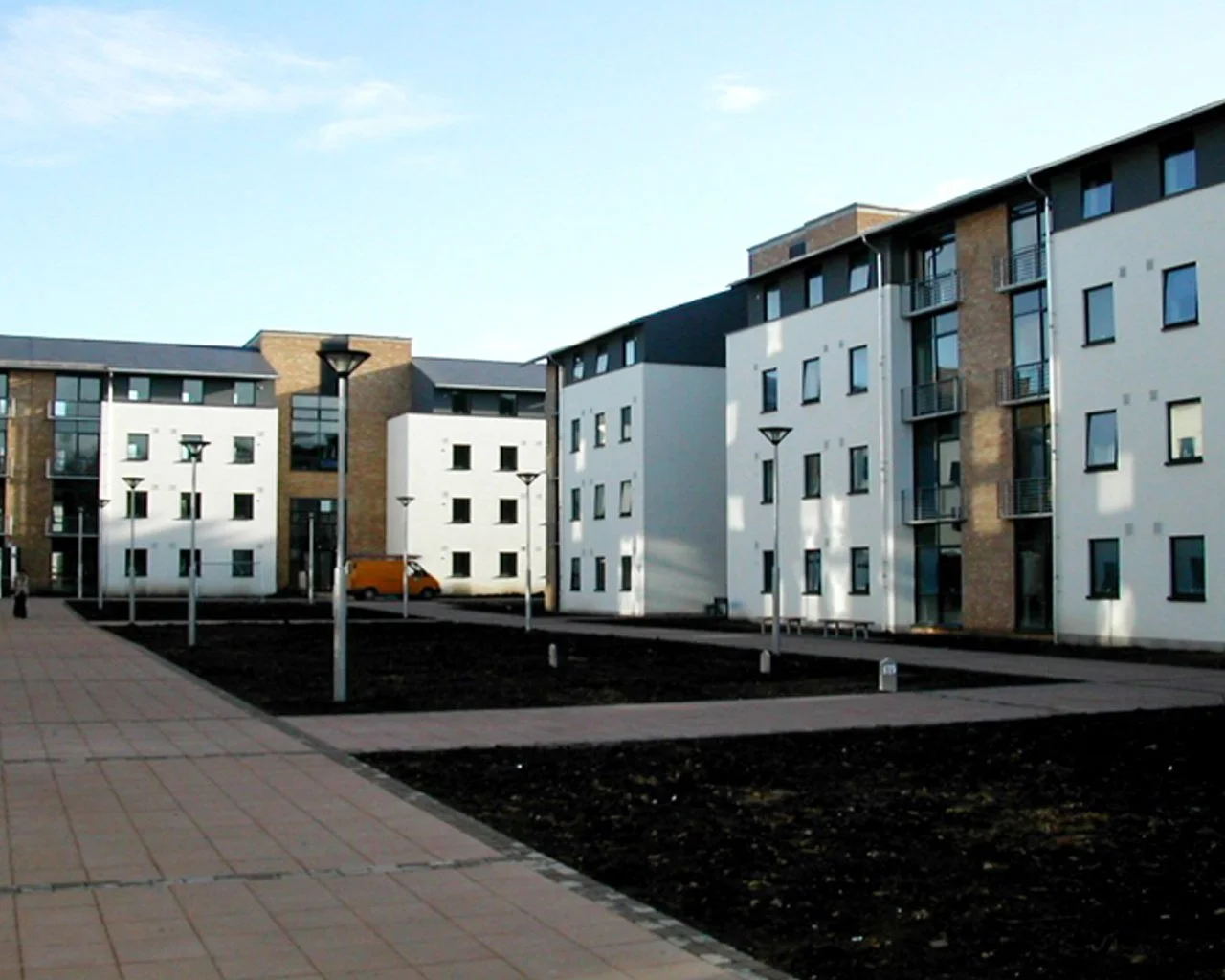 Modern apartment complex with white and brick facades, paved walkways, street lamps, and a yellow van parked outside during daytime.