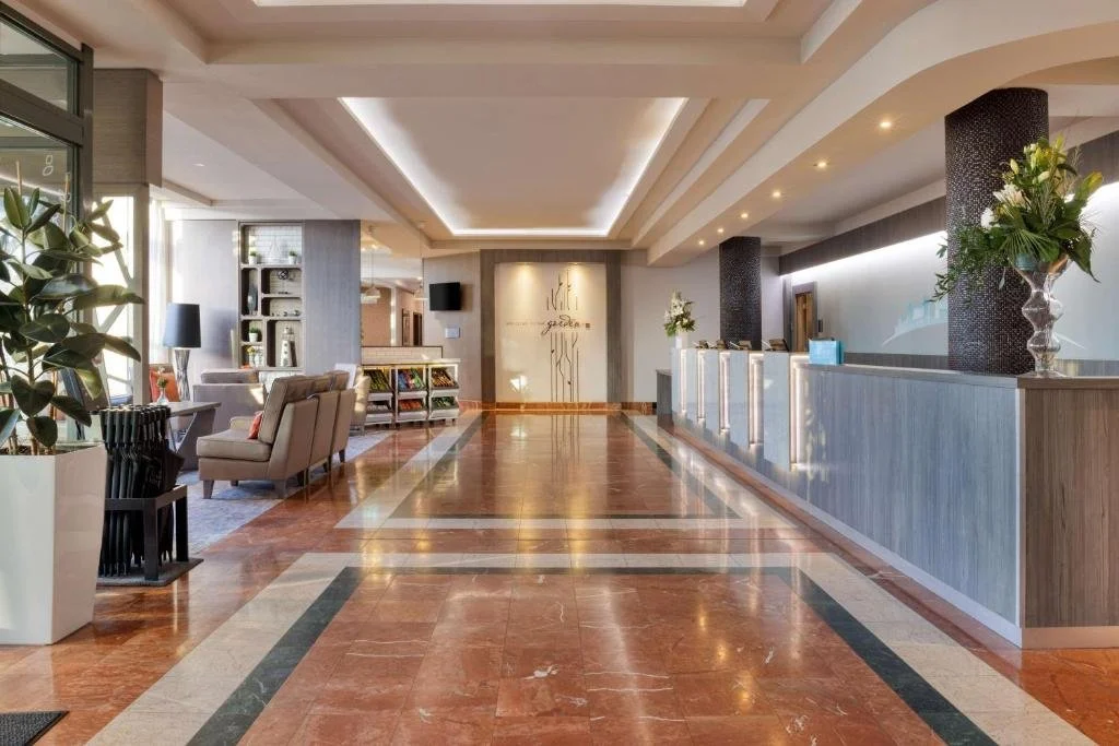 Hotel lobby with polished marble floors, a check-in desk, seating area with comfortable chairs and tables, decorative plants, and warm lighting.
