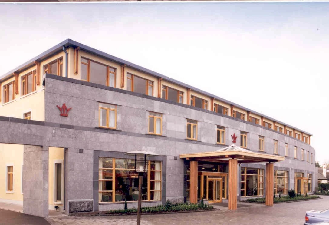 A modern, multi-story hotel building with a gray stone facade, wooden window frames, and a covered entrance with large wooden columns, featuring red logo symbols above the windows.
