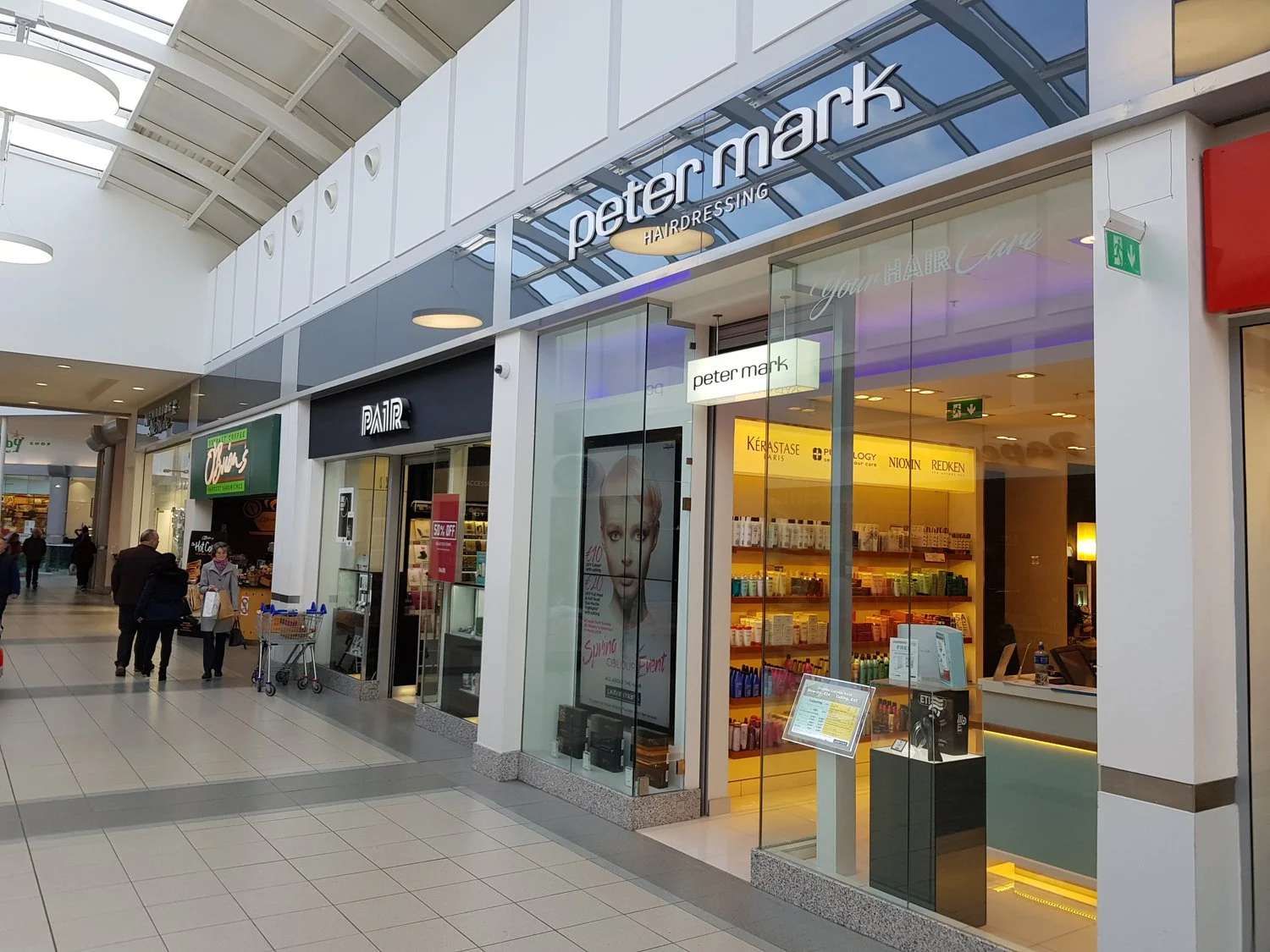 View of a shopping mall with a Peter Mark hair salon storefront, displaying products inside, and adjacent stores including a food vendor and a salon with a digital advertisement featuring a woman's face. Several shoppers are walking past.