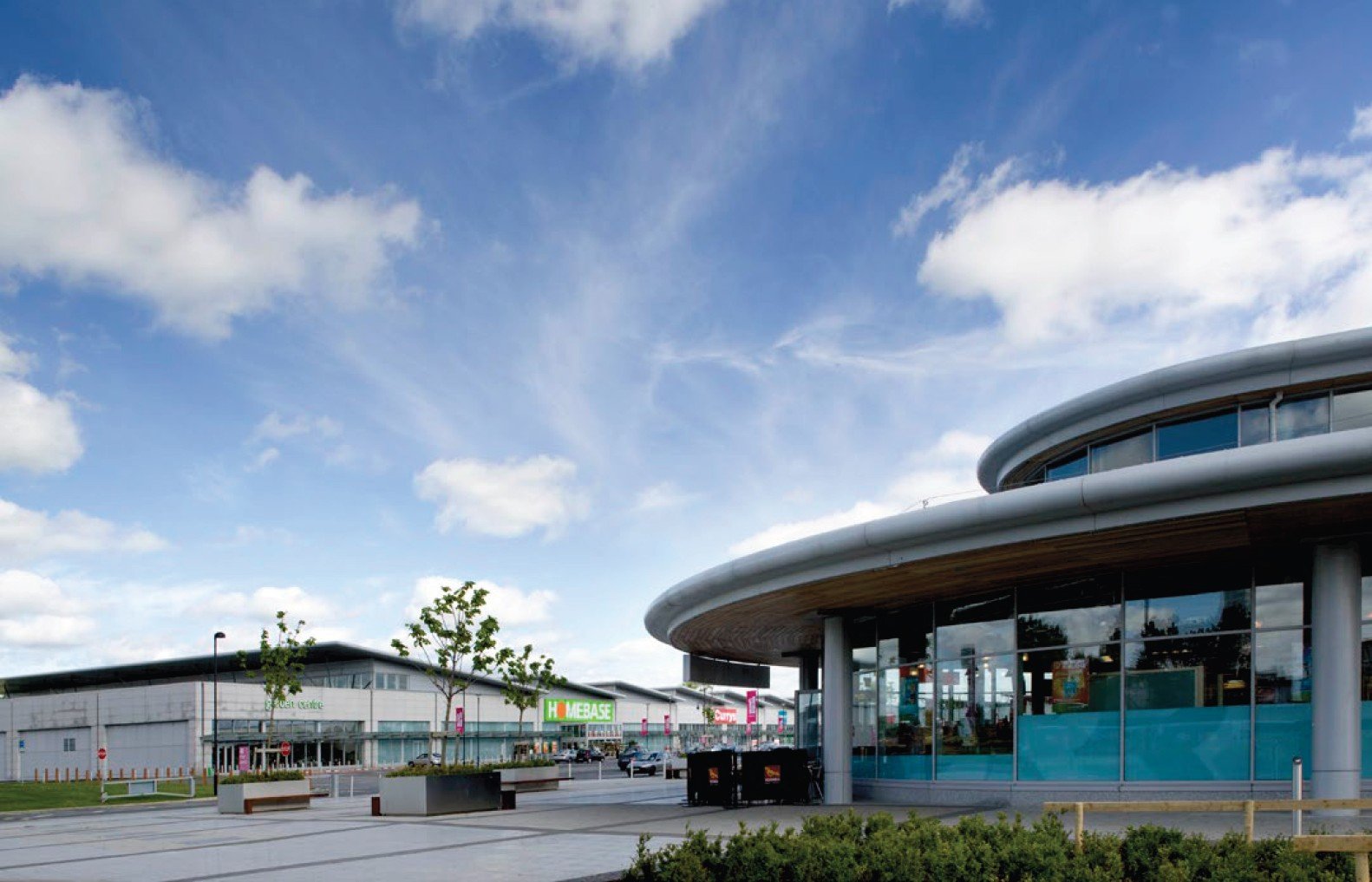 Exterior view of a modern shopping center with a Homebase store in the background, featuring a curved glass building, parking lot, and few trees under a partly cloudy sky.