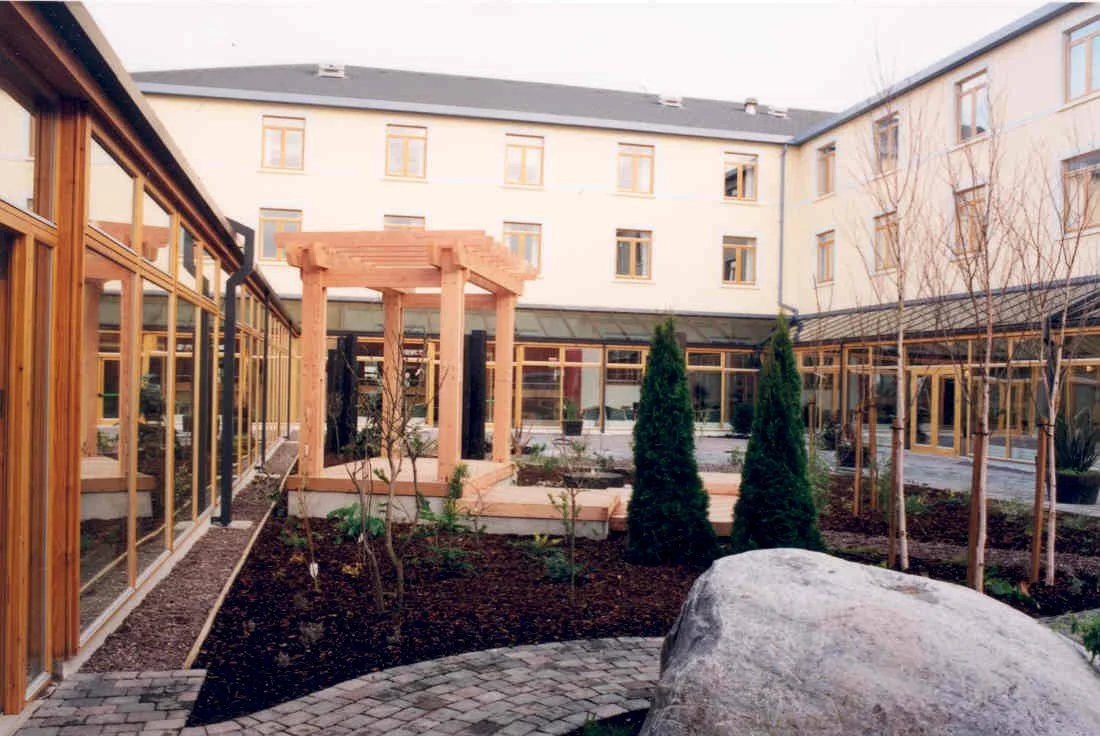 A courtyard garden surrounded by multi-story building with large windows, featuring a small wooden pergola, trees, and landscaped soil with paving stones.