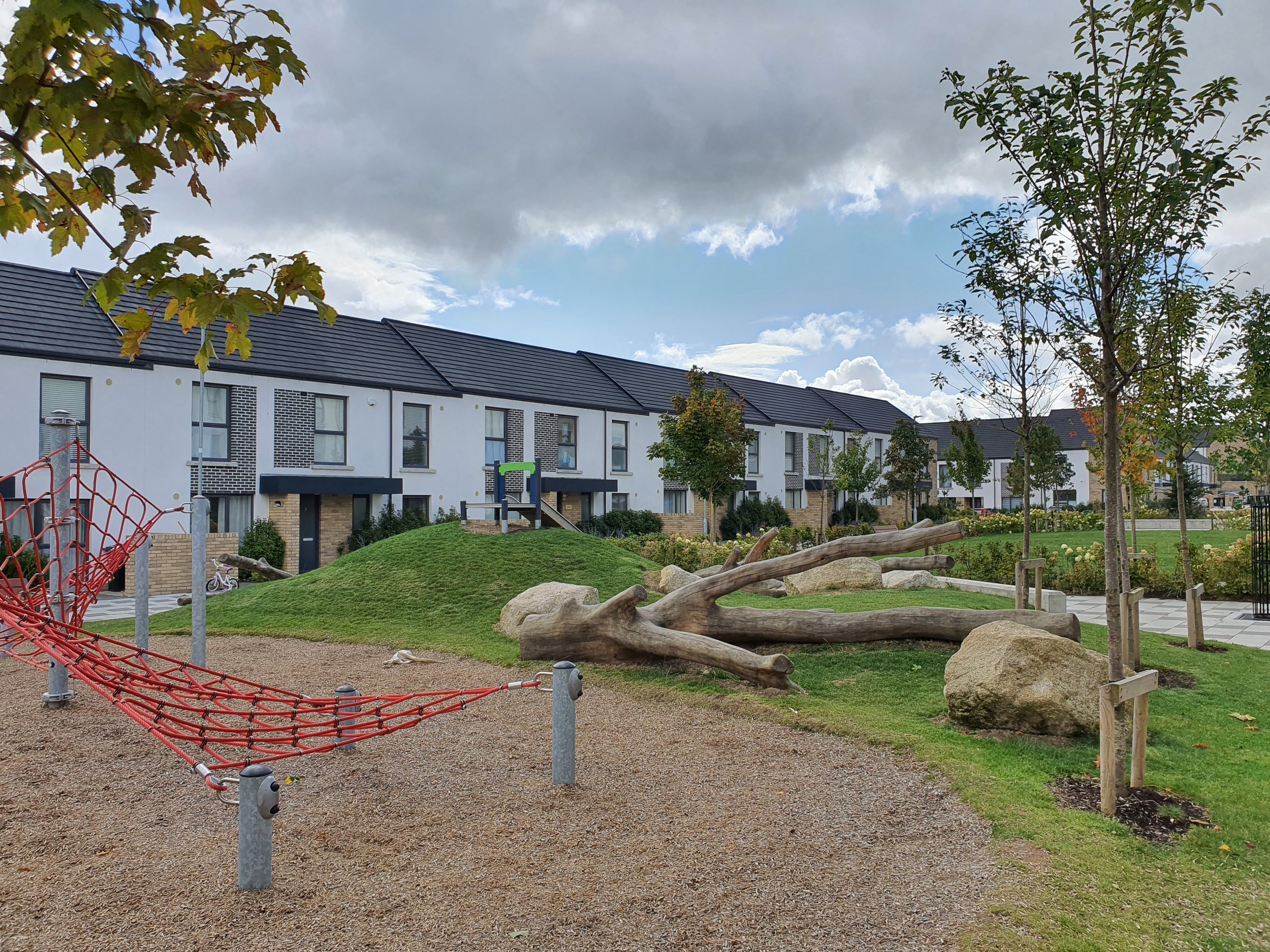 A residential neighborhood with modern townhouses, a small playground with a red climbing net, trees, rocks, and a grassy hill.