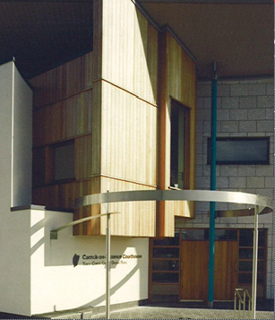 Exterior view of a modern building with wooden paneling, a white section, and large windows, featuring a curved metal canopy.