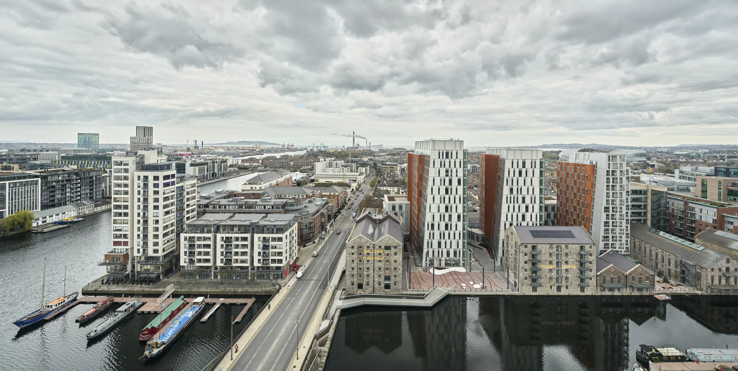 A cityscape featuring modern high-rise buildings along a river, with boats docked at a marina, and a bridge crossing the river under a cloudy sky.