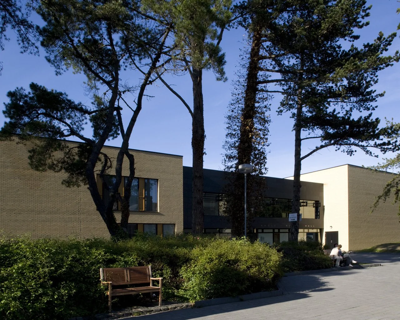 A modern building with large windows, surrounded by tall trees and greenery. There are two people sitting on a bench near the building and a wooden bench in the foreground.