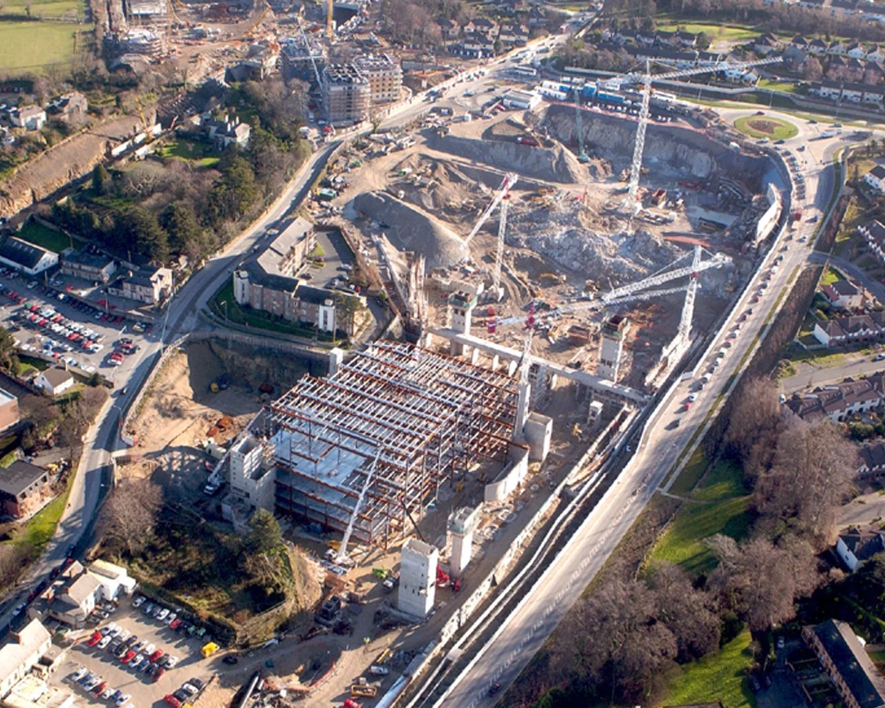 An aerial view of a construction site featuring a building under construction with exposed steel framework, several cranes, and surrounding residential and commercial buildings.