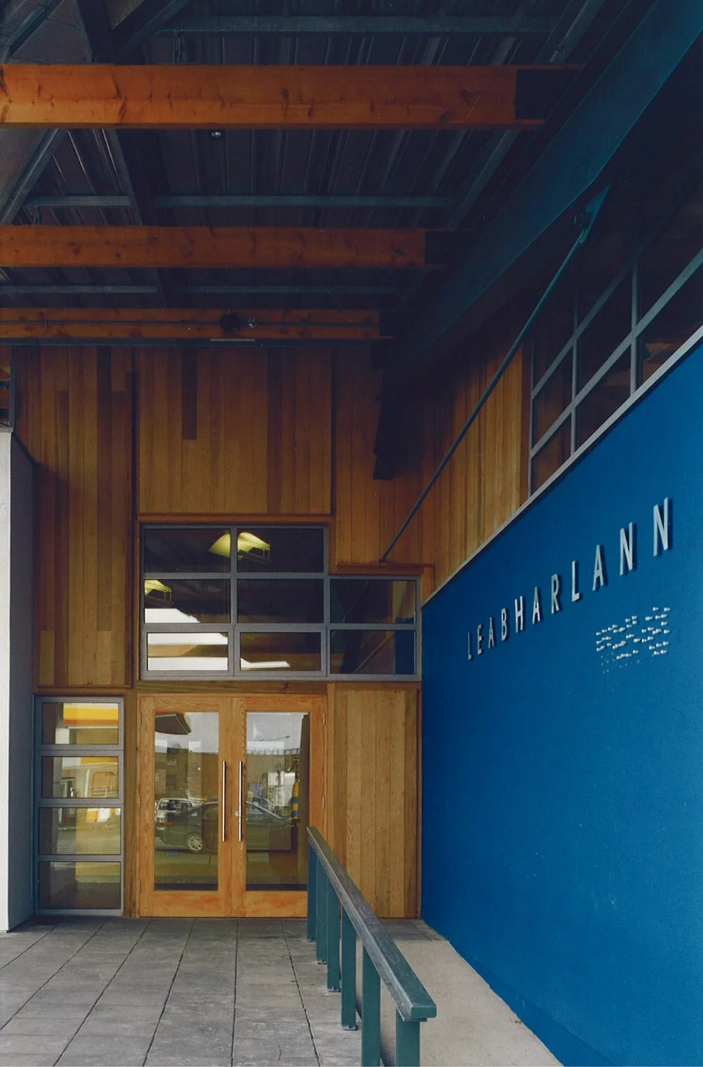 Entrance with wooden and blue wall, sign reading 'LEABHARLANN', glass double doors, railing, and parking lot reflected in the glass.