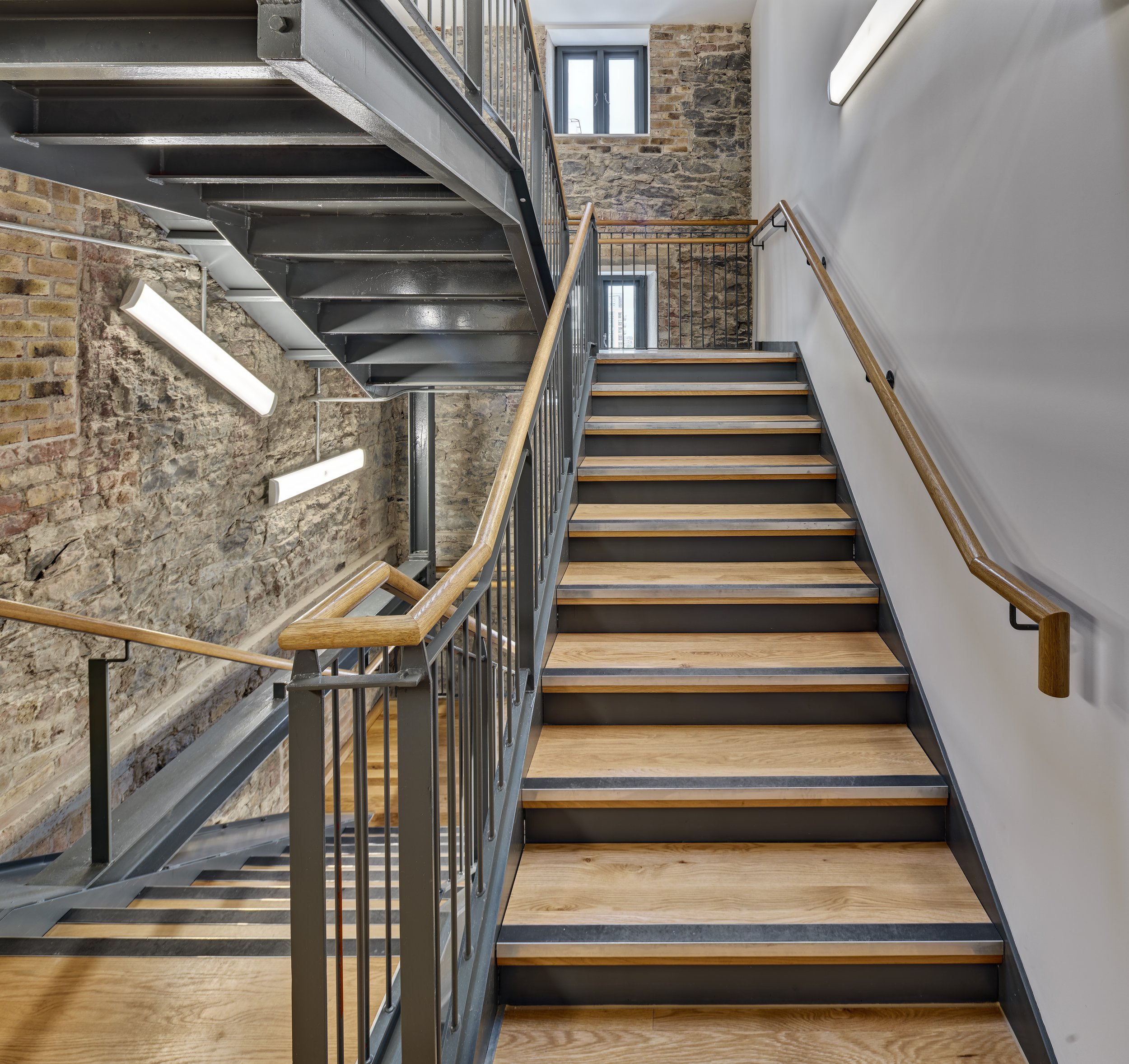Modern indoor staircase with wooden handrails, metal railings, wooden steps with black trim, exposed brick wall, and white wall with linear light fixtures.