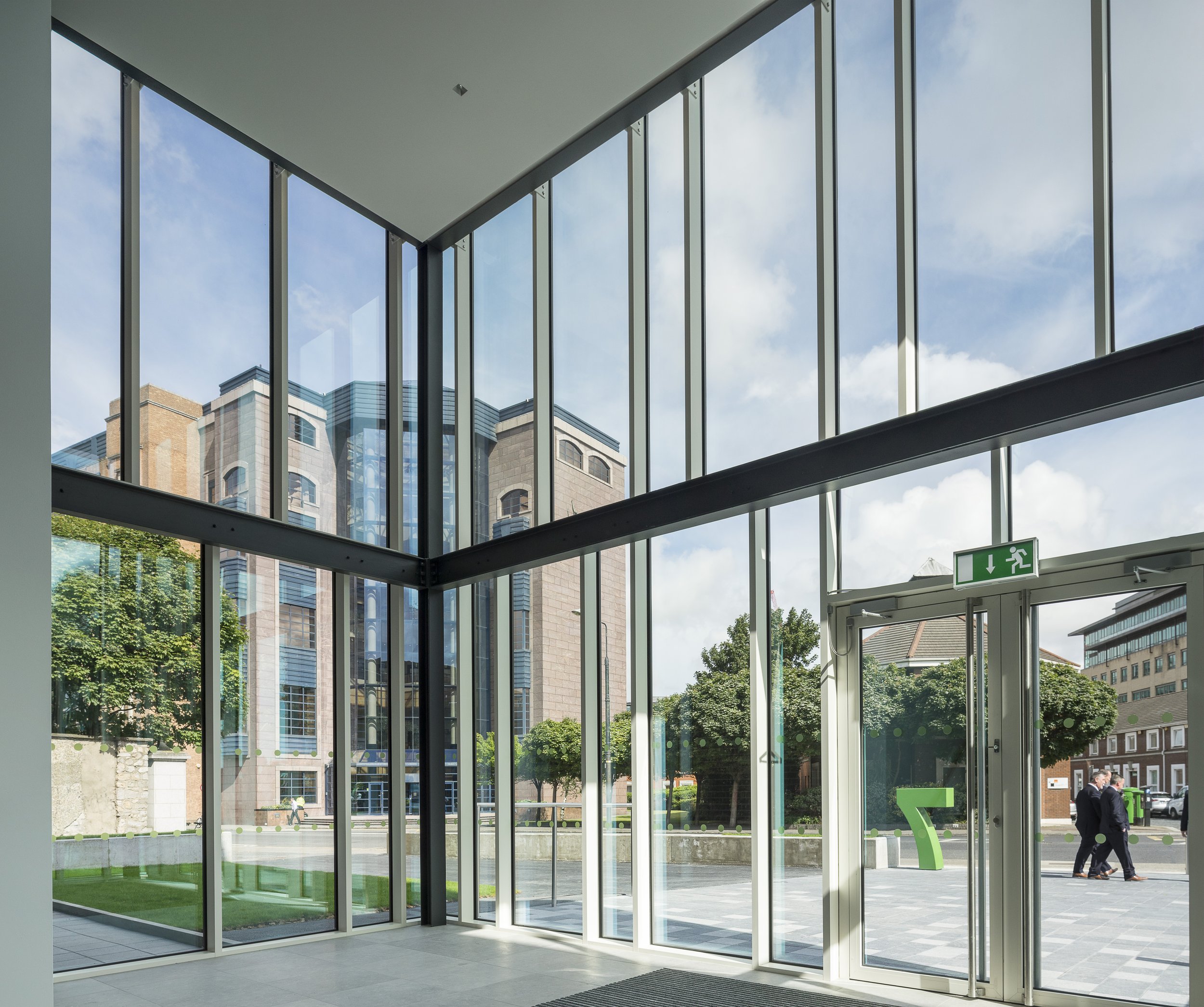 Interior view of a modern building with large glass windows and door. Outside, there are trees, a few people walking, and a large green number 7 on the sidewalk. The sky is partly cloudy.