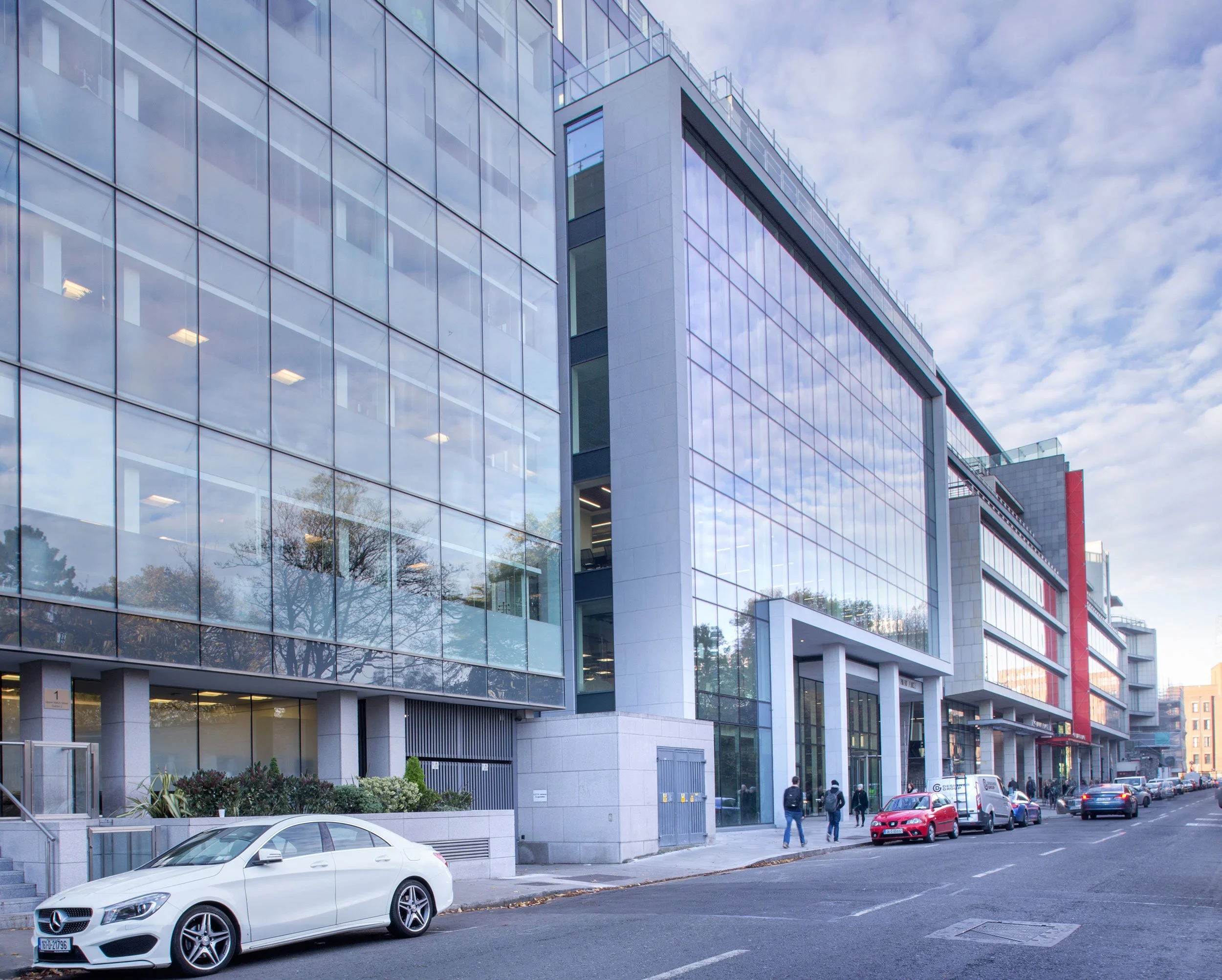 Modern glass-fronted office building with reflections of trees and the sky, parked cars, and pedestrians walking along the sidewalk.