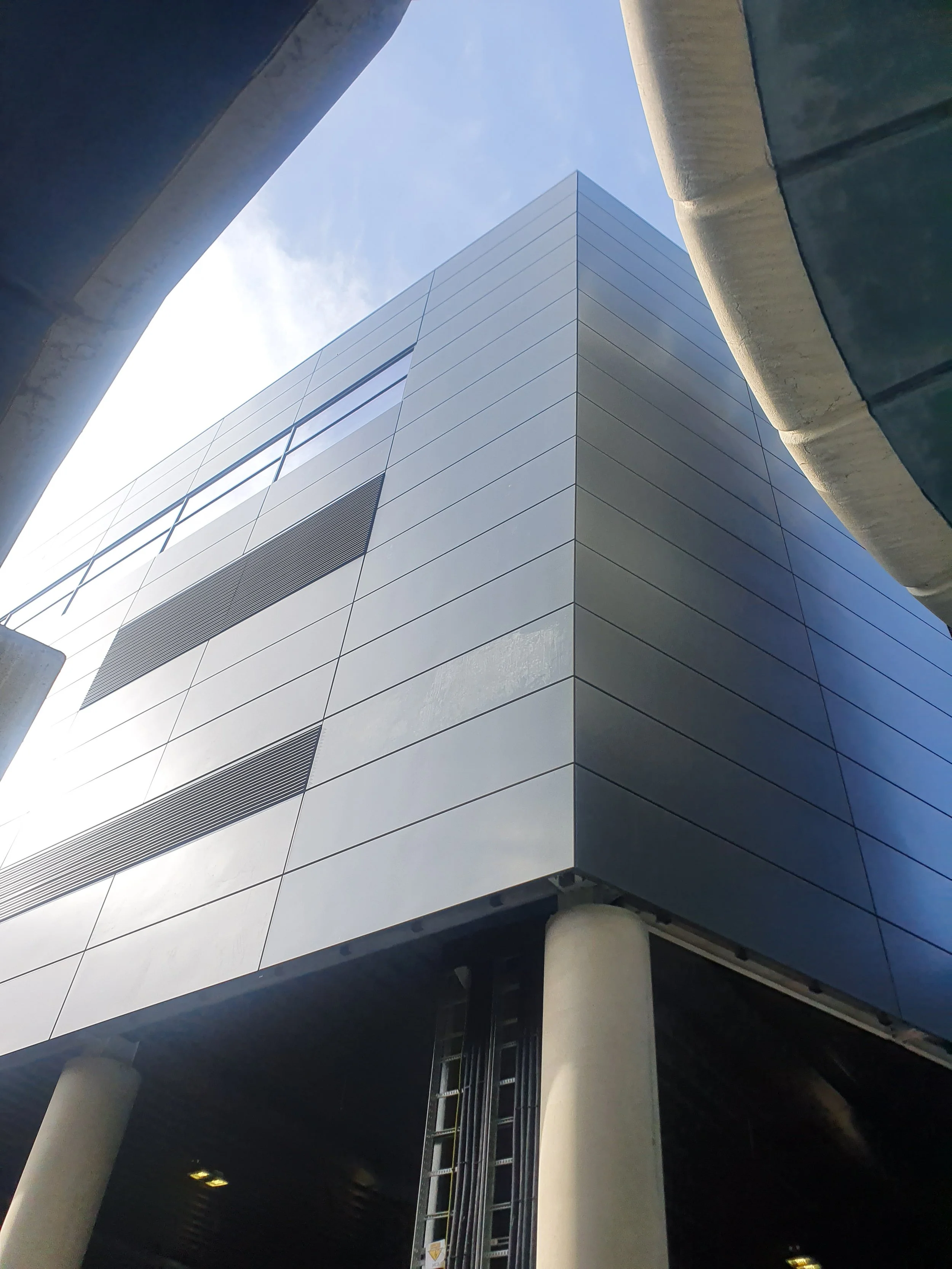Low-angle view of a modern building with sleek blue and gray panels, supported by concrete columns, against a blue sky with faint clouds.