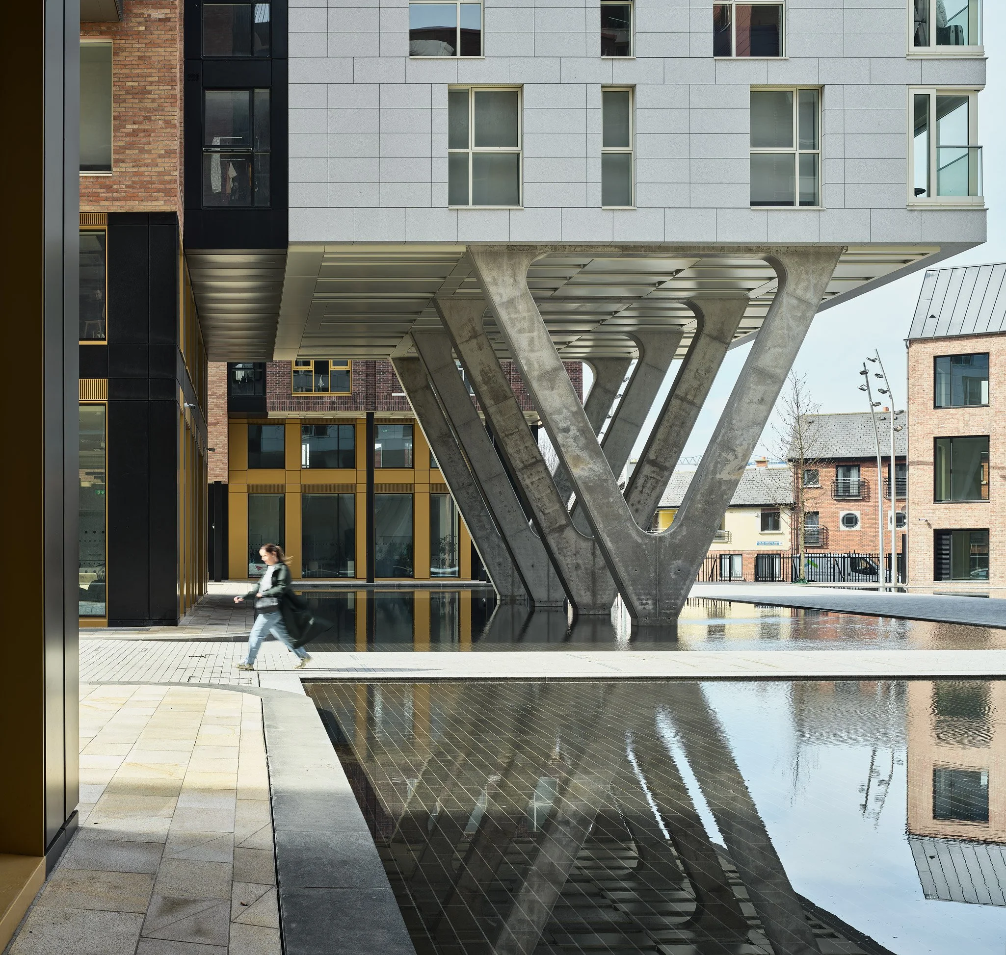 Modern urban scene featuring a building with a unique V-shaped concrete support structure, glass windows, and a reflective water feature mirroring the architecture. A woman wearing a mask walks across the paved area.