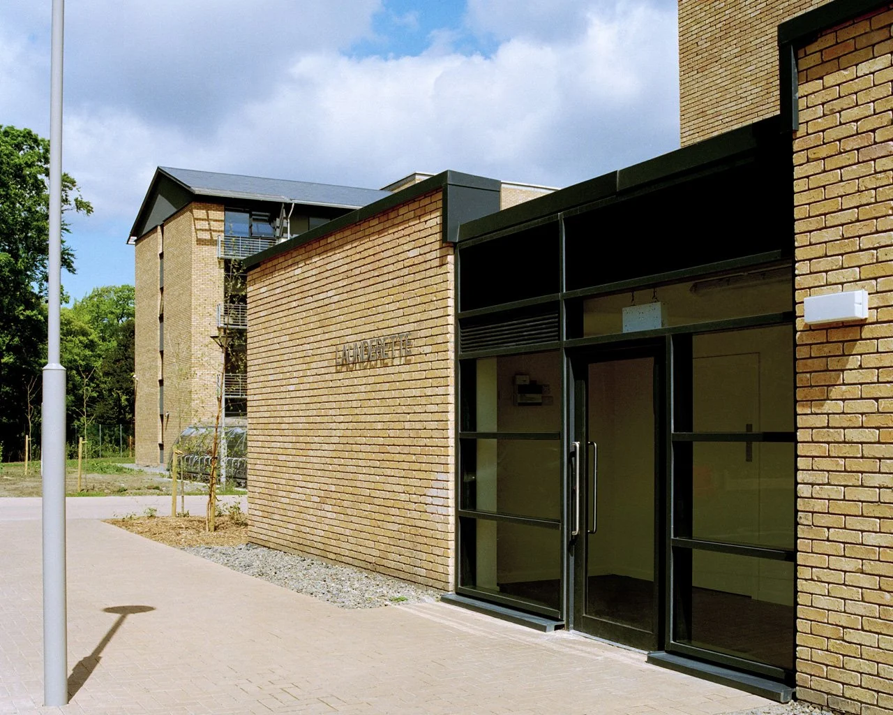 Exterior view of a modern brick building with glass entrance door, sign reads 'Andersfeld,' some trees, and a blue sky with clouds.