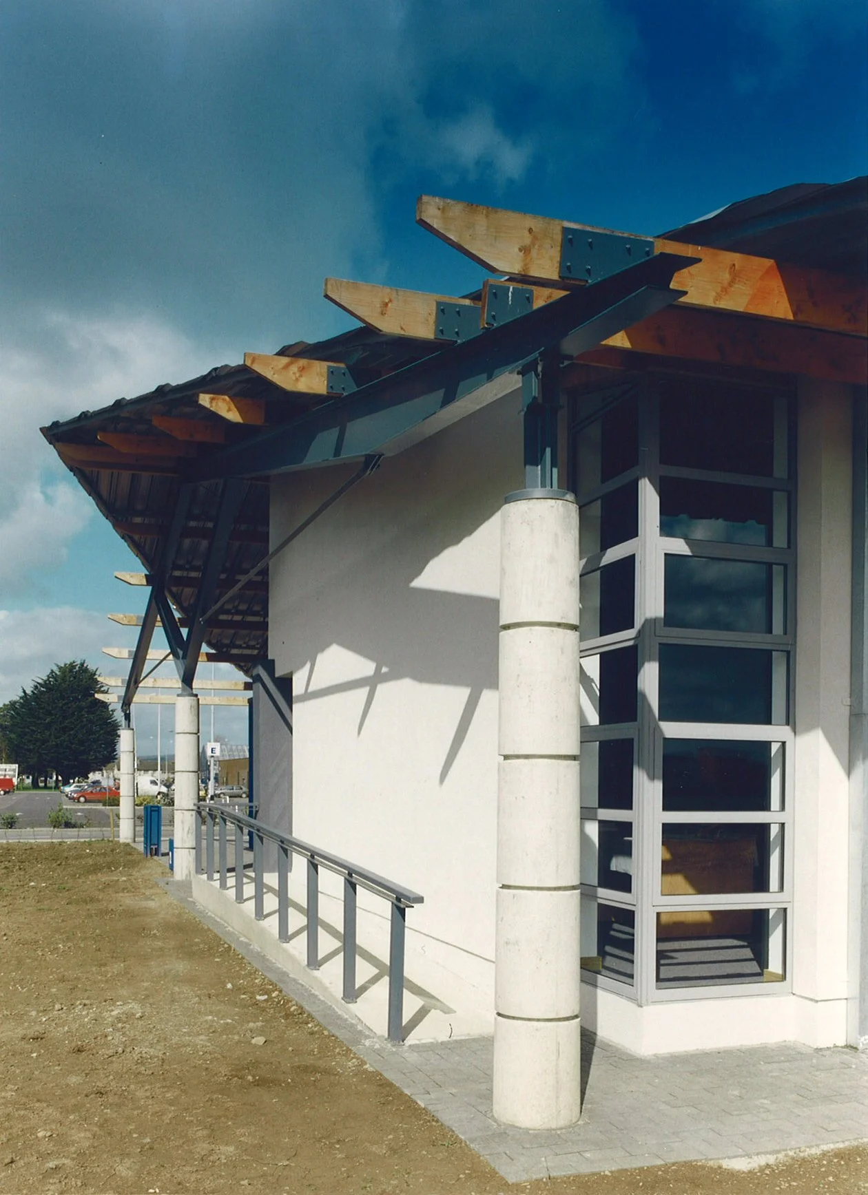 A modern building under construction showing exposed wooden beams on the roof and a large corner window with gray framing, supported by a concrete pillar, with a metal ramp and a dirt area in the foreground.