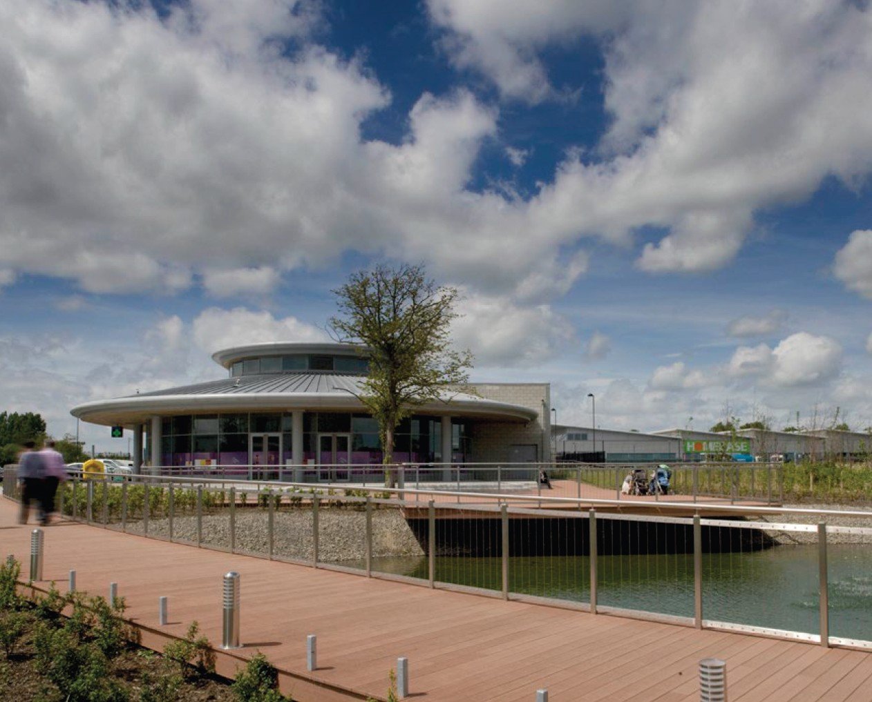 Modern building with circular design, glass windows, and a metal roof, situated by a waterway with a boardwalk and a tree in the foreground under a partly cloudy sky.