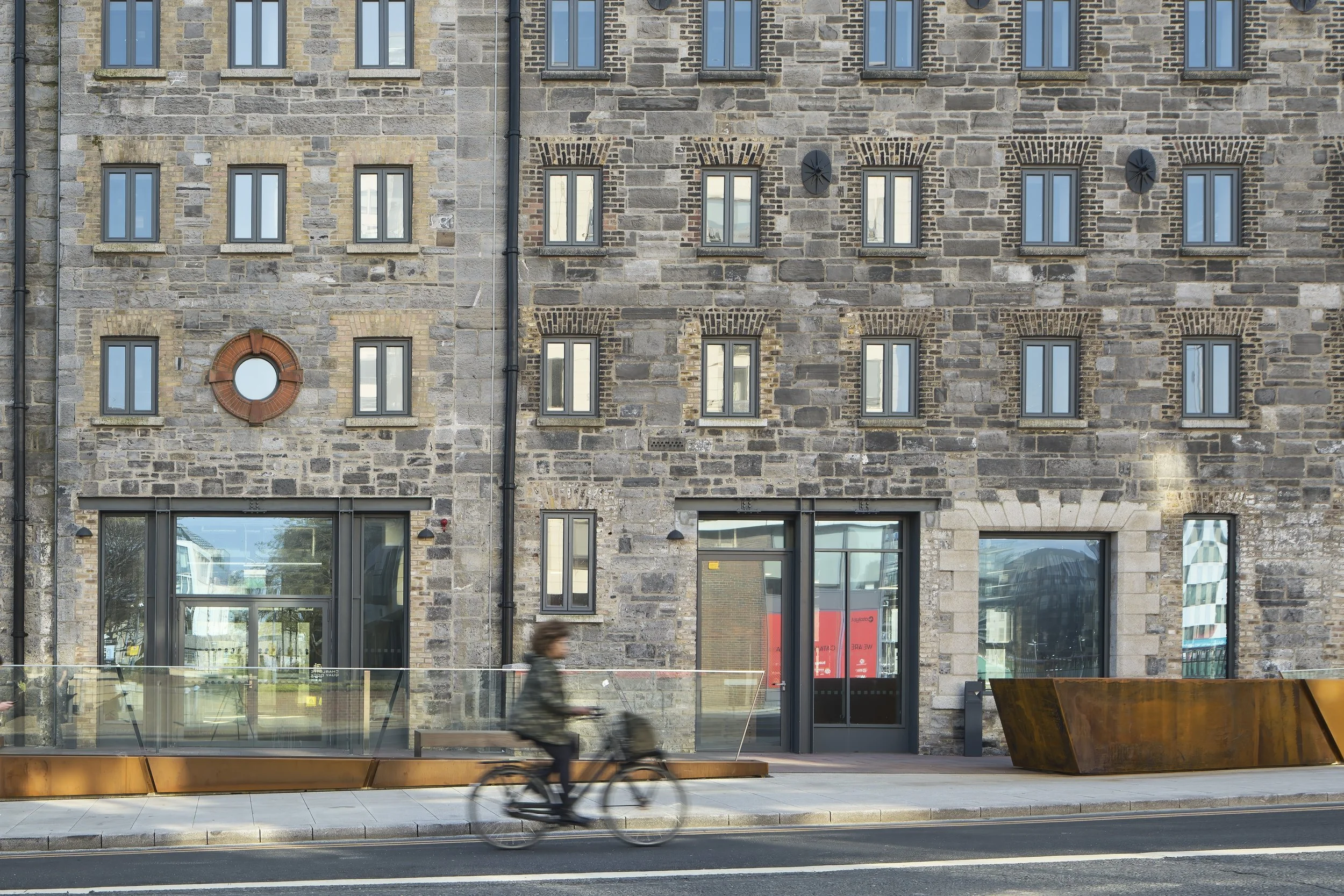 Person riding a bicycle on the street in front of a stone building with multiple windows and glass entrance doors.