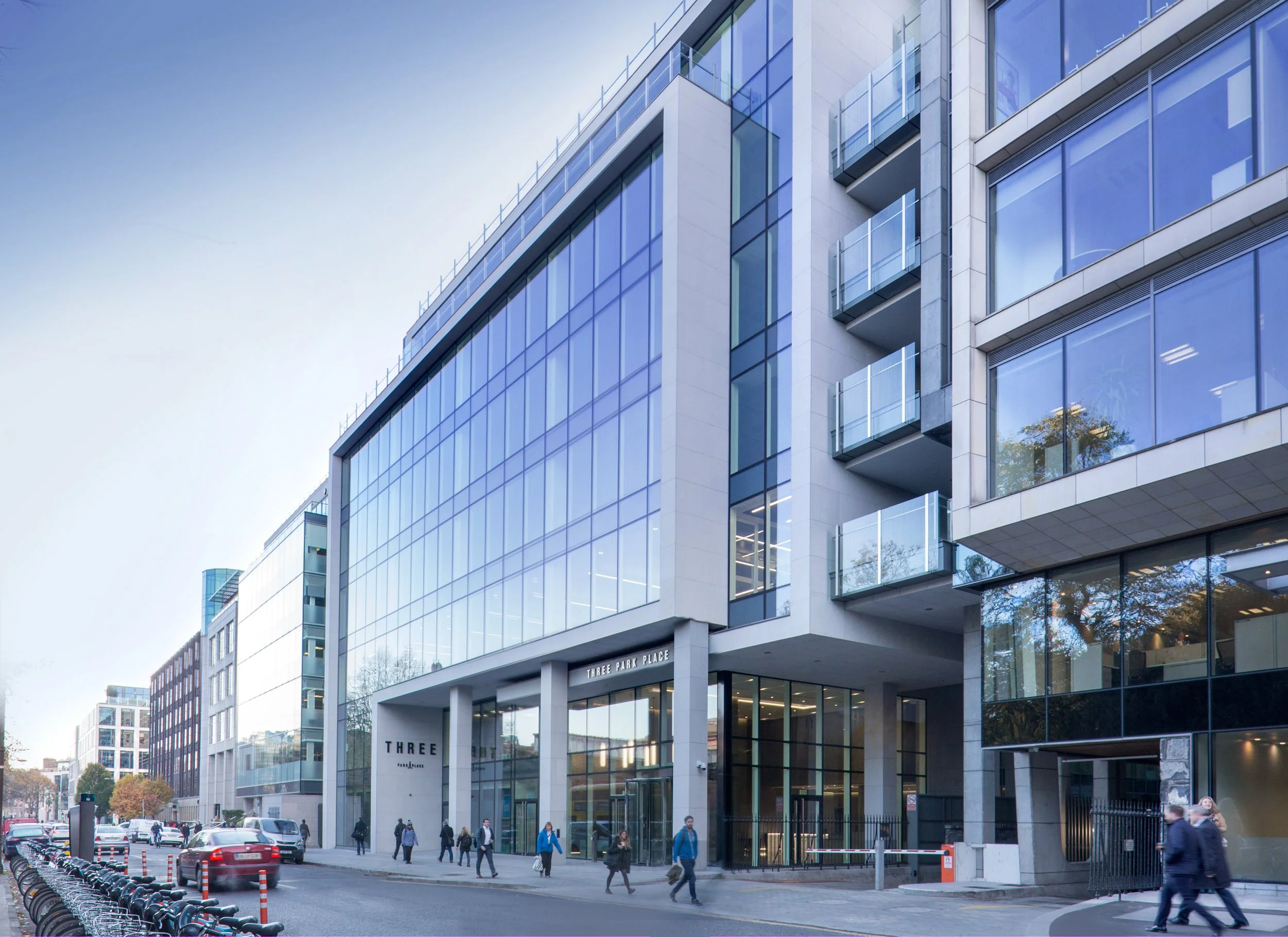 Modern multi-story office building with glass windows and balconies, people walking on the sidewalk, cars parked along the street, and the name 'Three Park Place' on the building entrance.