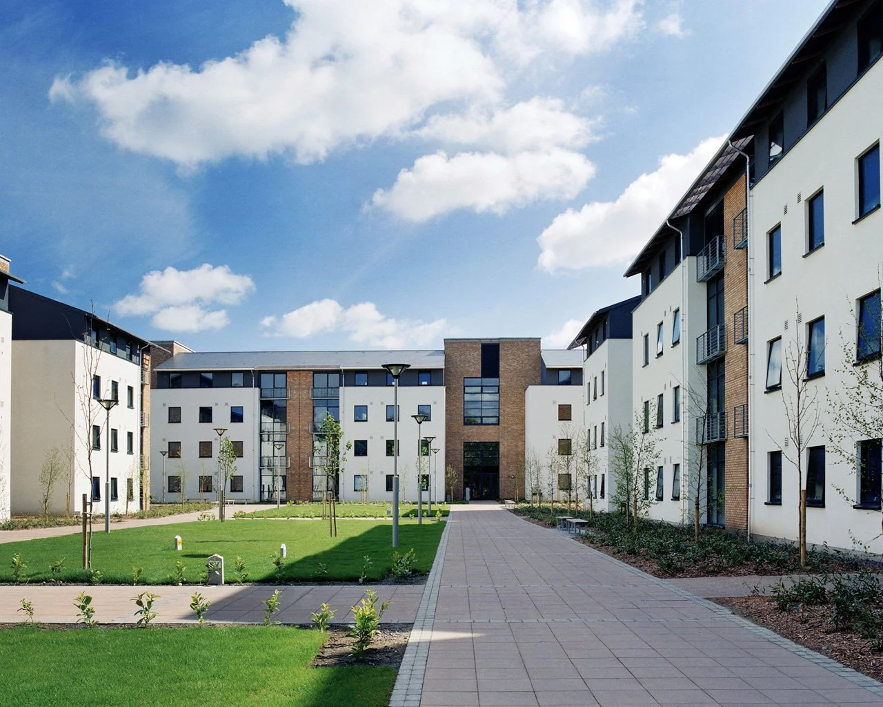 View of modern residential apartment complex with landscaped green courtyard, paved walkways, young trees, and multiple-lamp posts under a partly cloudy sky.
