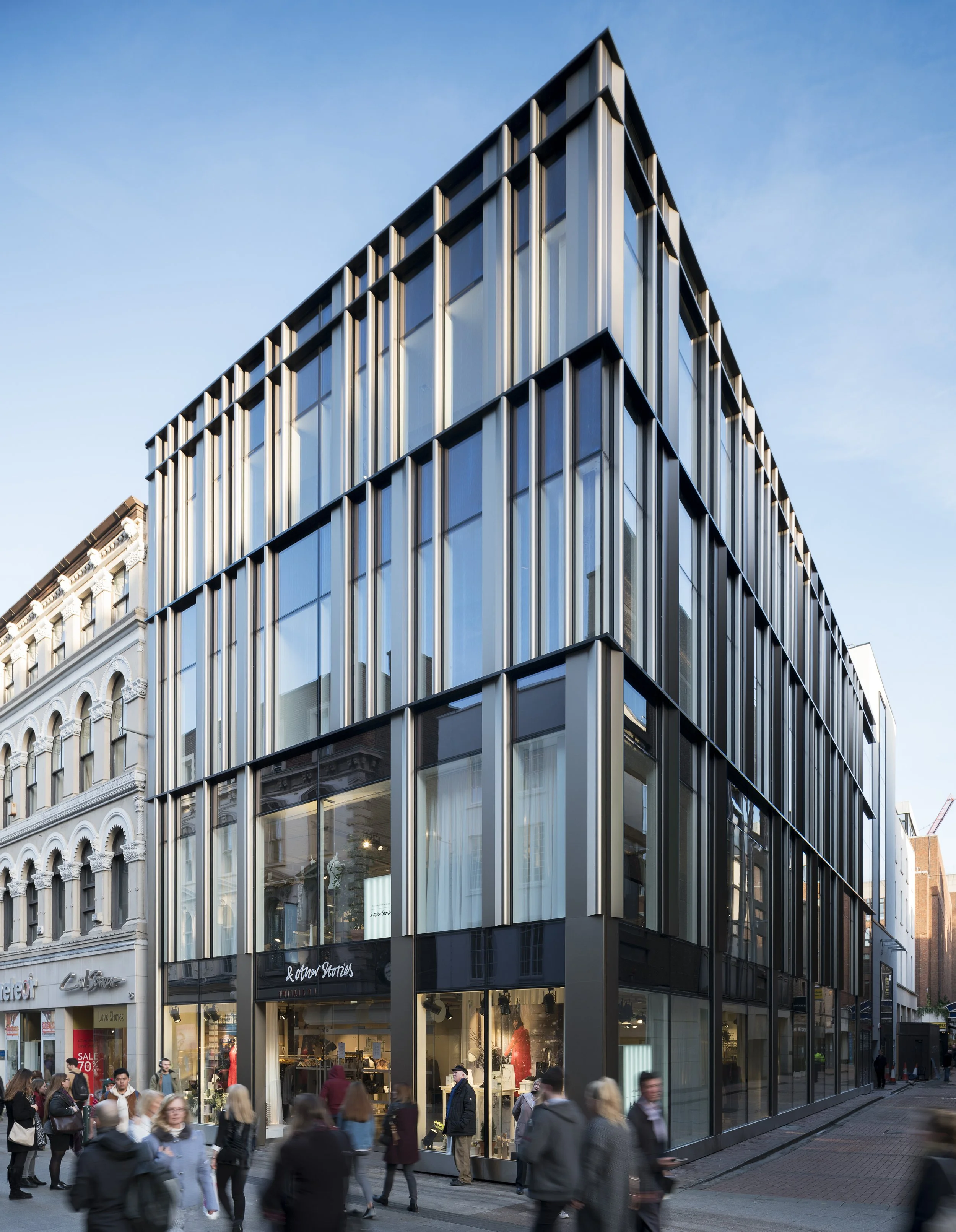 Modern multi-story glass building with vertical metal accents on a city street, with pedestrians walking by.