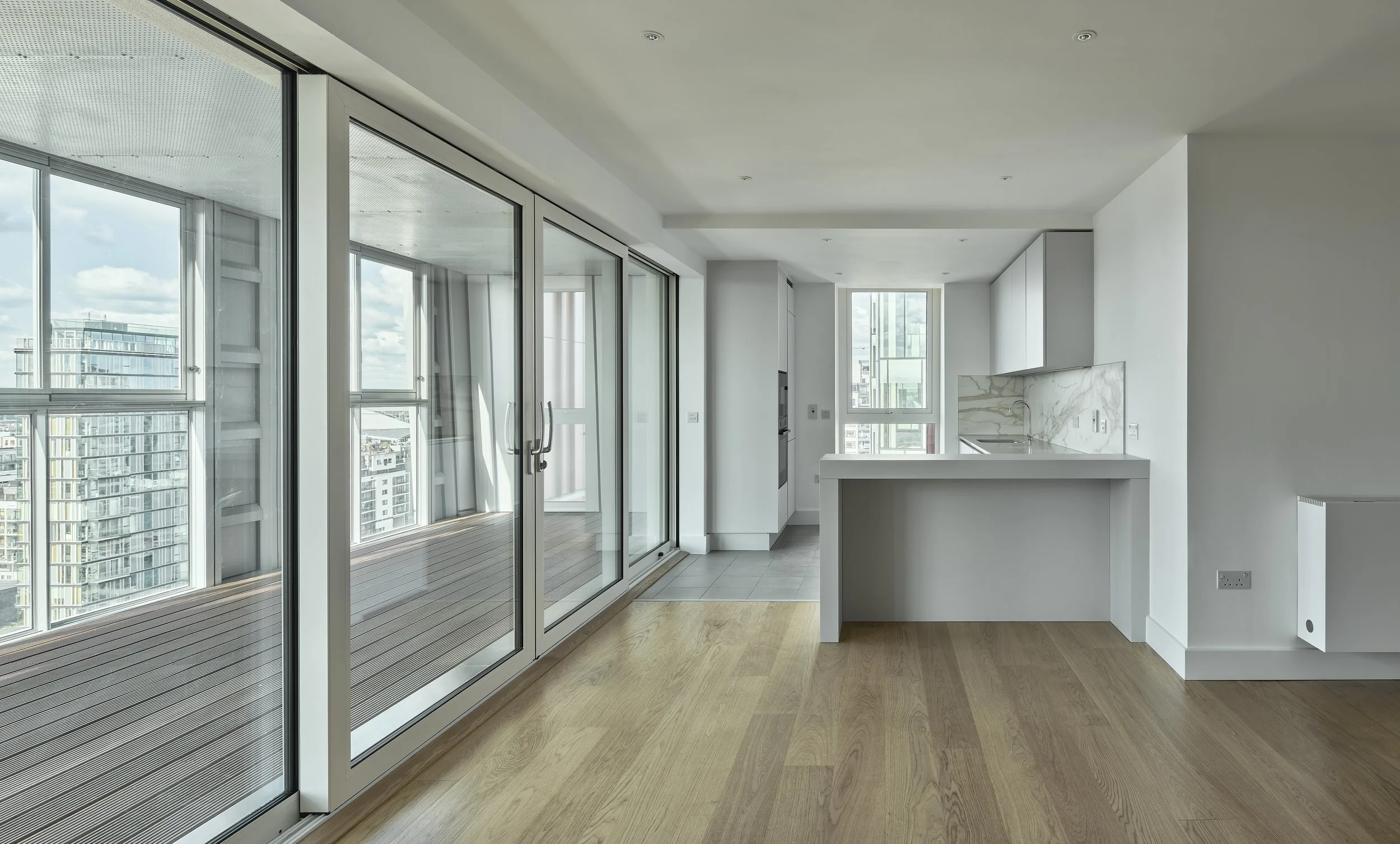 Modern apartment interior with large glass sliding doors opening to a balcony, a white kitchen island with marble backsplash, and hardwood flooring.