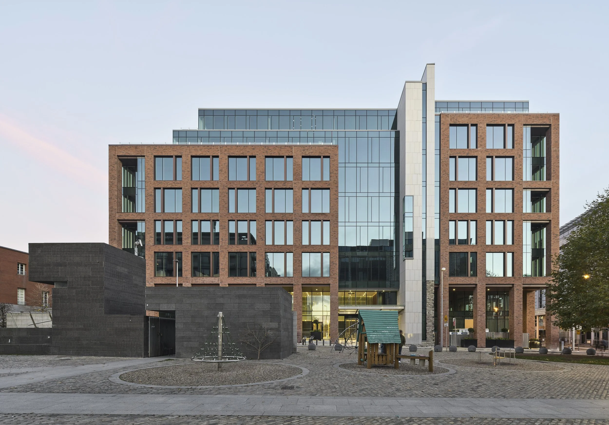 Modern multi-story building with brick and glass facade, surrounded by a small playground area with a slide, trees, and cobblestone pavement.