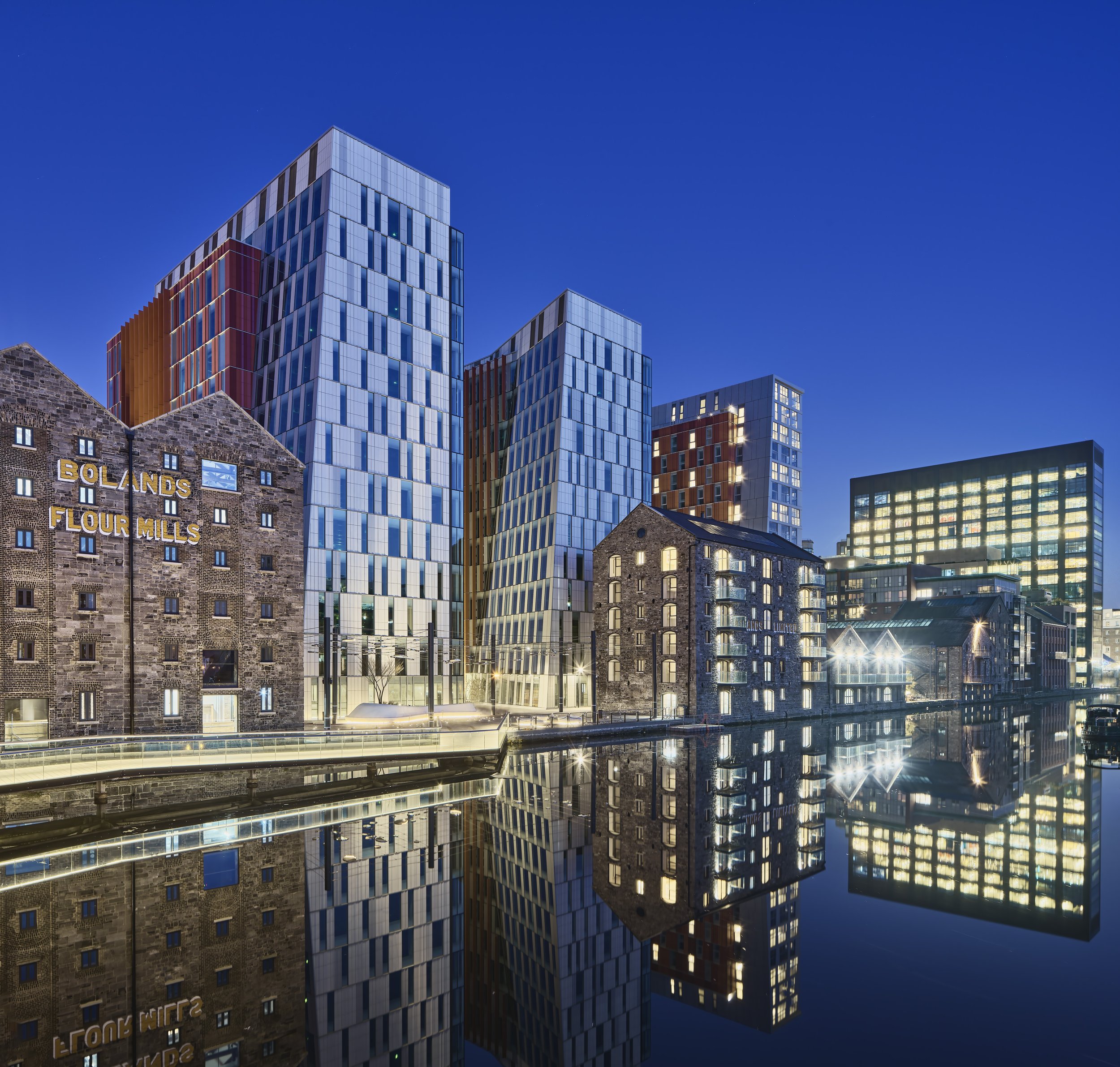 Night view of modern city buildings reflecting in a water canal, with a historic stone building on the left titled 'BOLAND'S FLOUR MILLS'.