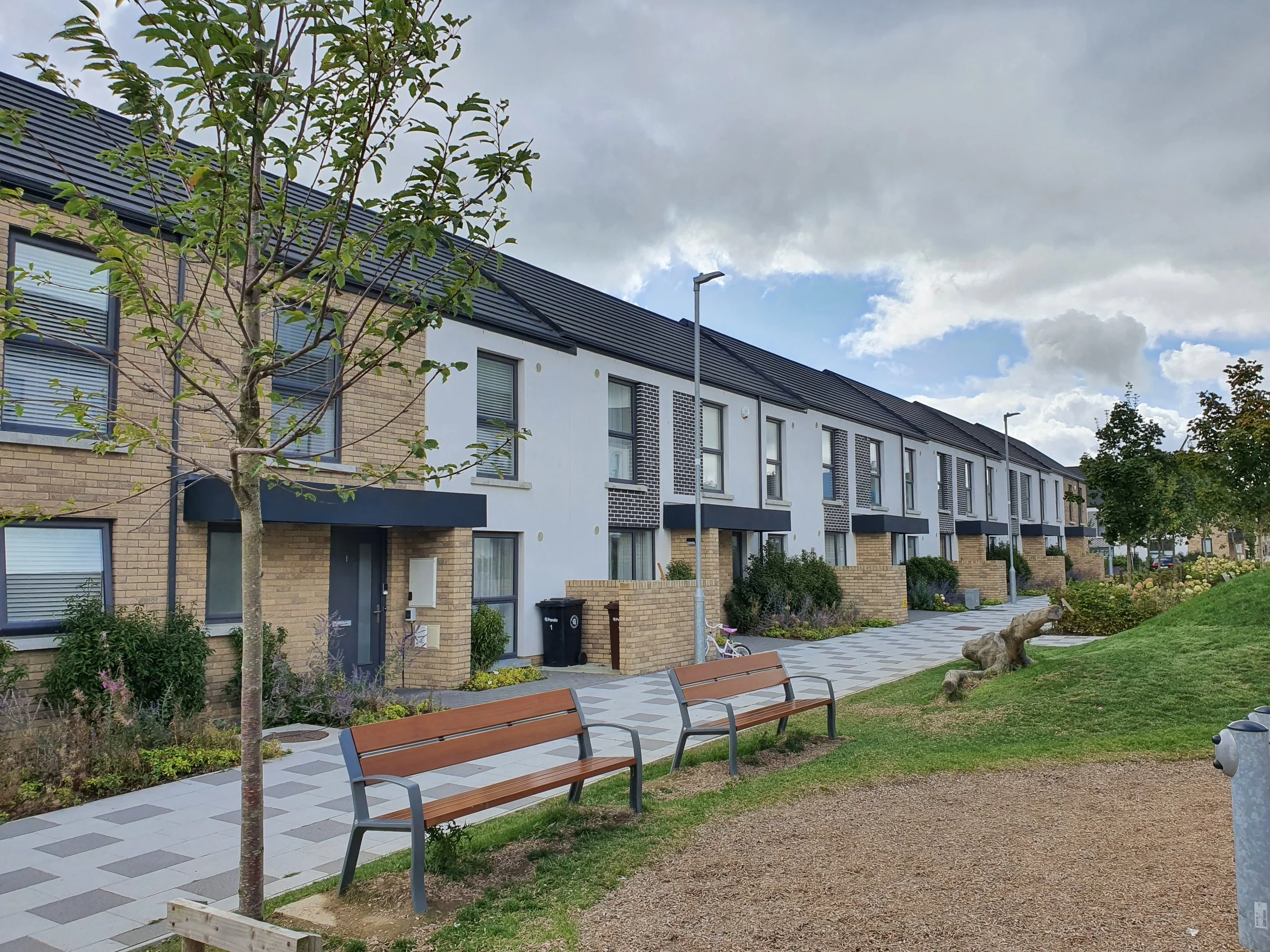 View of a modern residential apartment complex with multiple townhouses featuring brick and white exteriors, large windows with blinds, and black awnings. In the foreground, there are benches, small trees, a landscaped walkway, and a grassy area with rocks. The sky is partly cloudy.