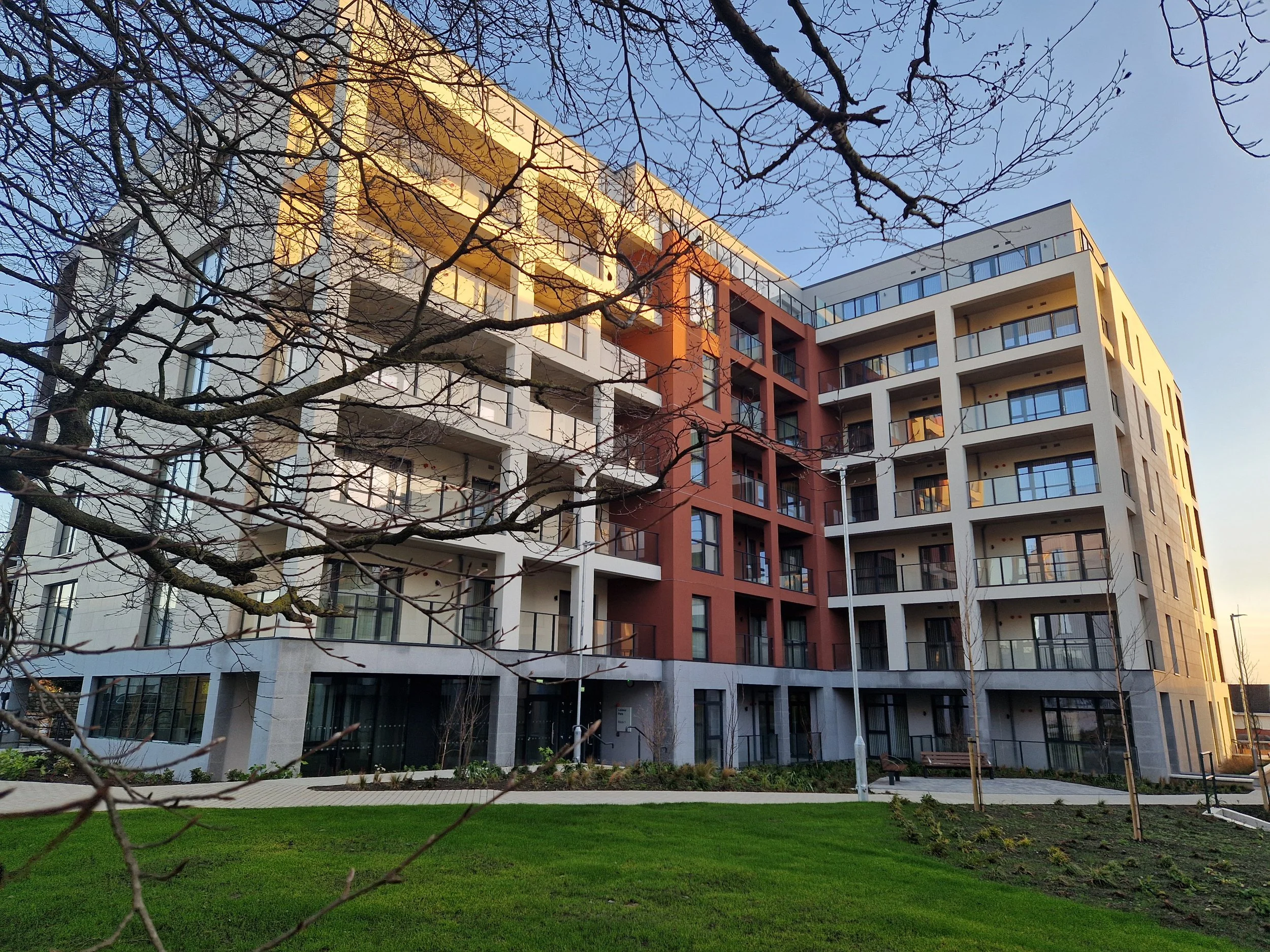Modern multi-story apartment building with balconies, set against a clear sky, surrounded by a well-maintained lawn and trees with leafless branches.