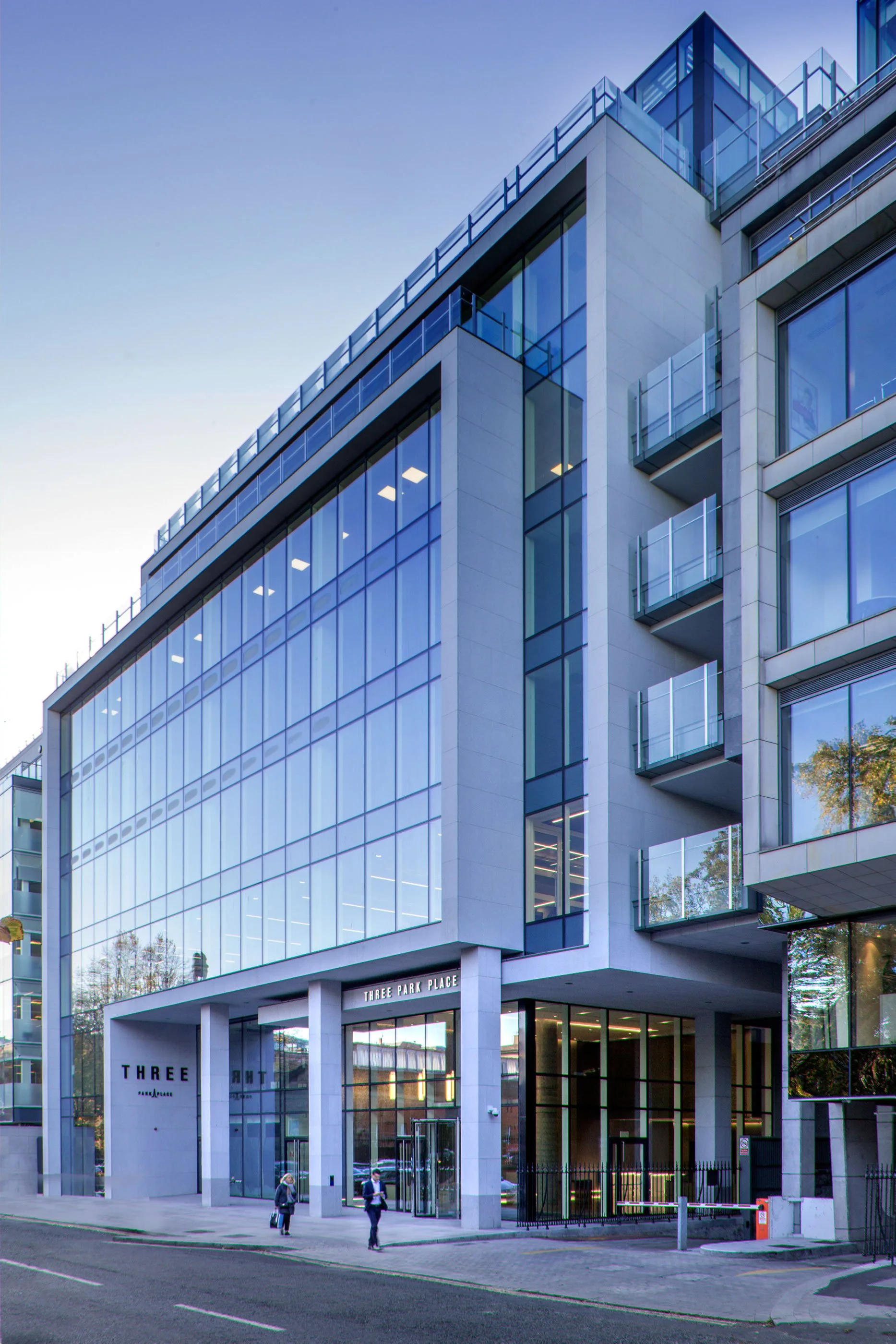 Modern multi-story glass office building with balconies, labeled 'Three Park Place', with pedestrians walking on the sidewalk in front.