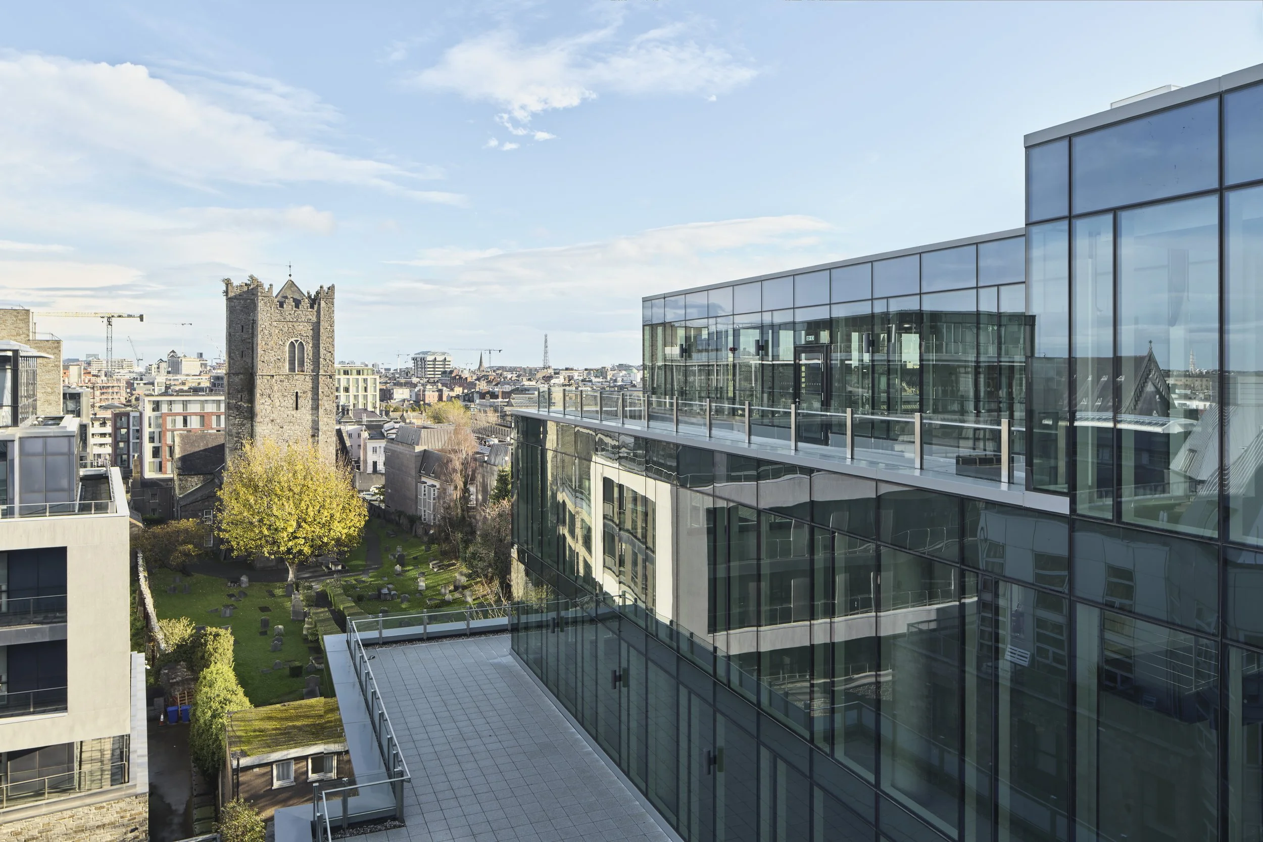 View of a modern glass building with a rooftop balcony, overlooking a historic stone church tower and surrounding cityscape with construction cranes and a clear blue sky.