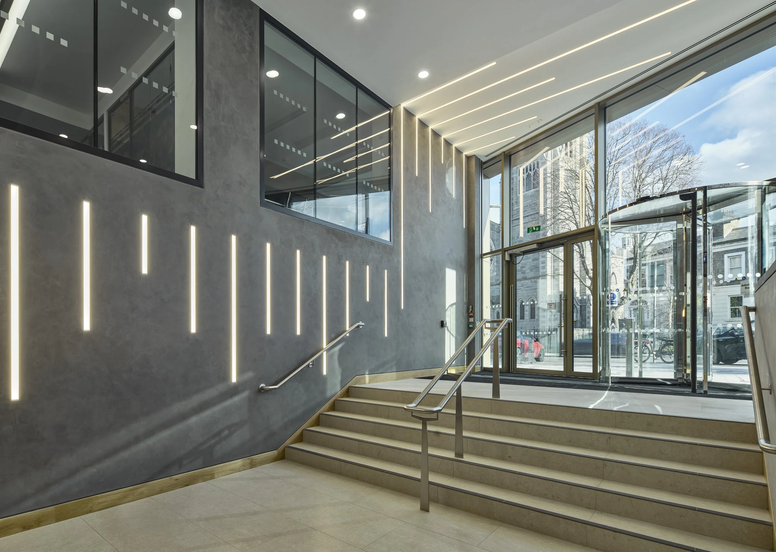 Modern building lobby with gray textured walls, vertical LED strip lighting, glass wall partitions, and large glass entrance doors with a view of city street outside, including cars and bicycles.