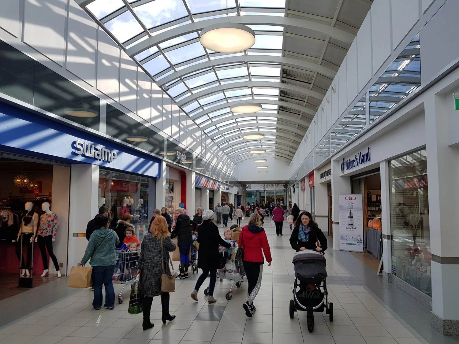 Shoppers walking through an indoor shopping mall with stores on either side and a glass ceiling above.