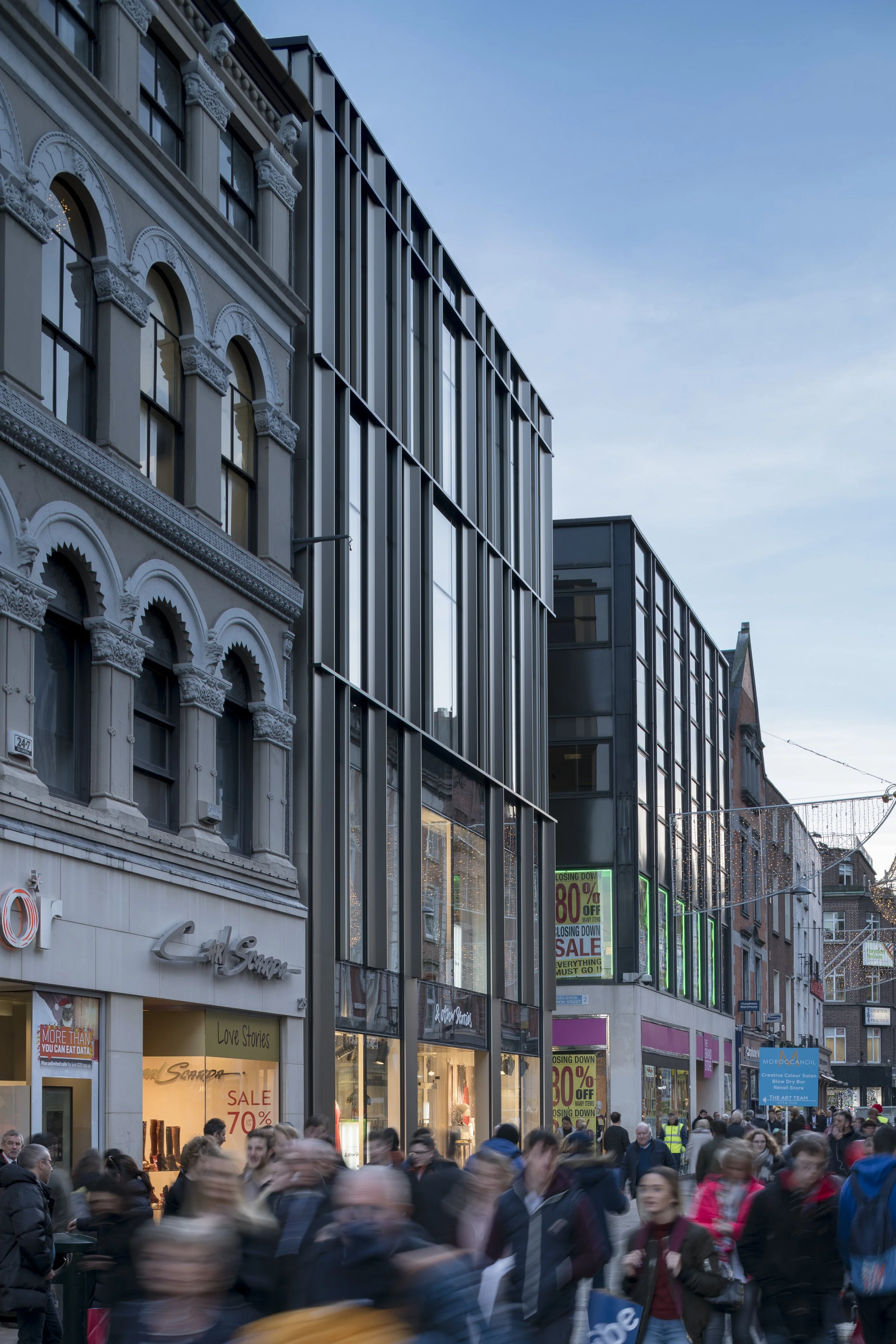 Busy city street with modern glass building and historic architecture, crowd of pedestrians in motion, neon signs, and sale advertisements.