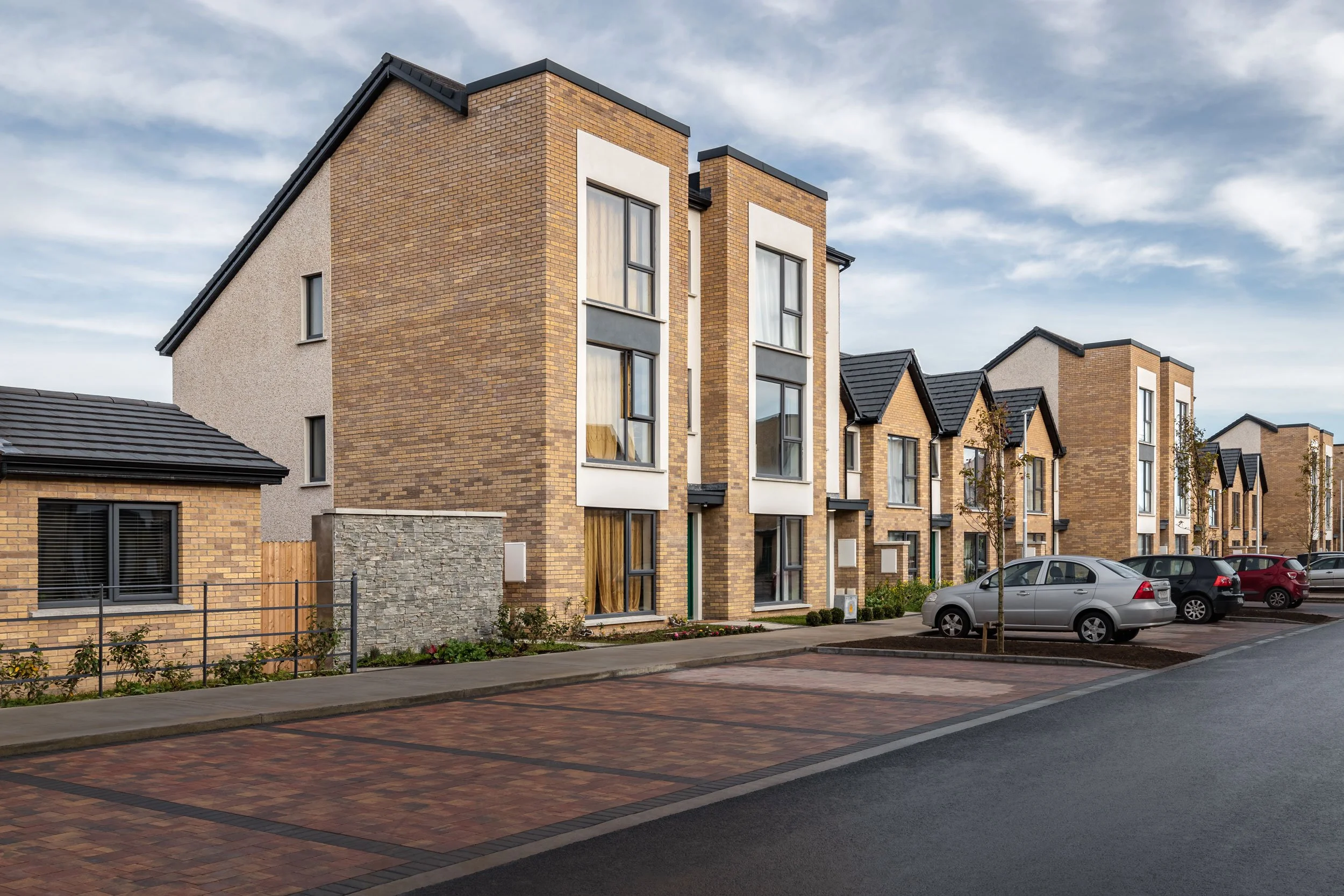 A row of modern brick townhouses with large windows, cars parked on the street, and a cloudy sky overhead.