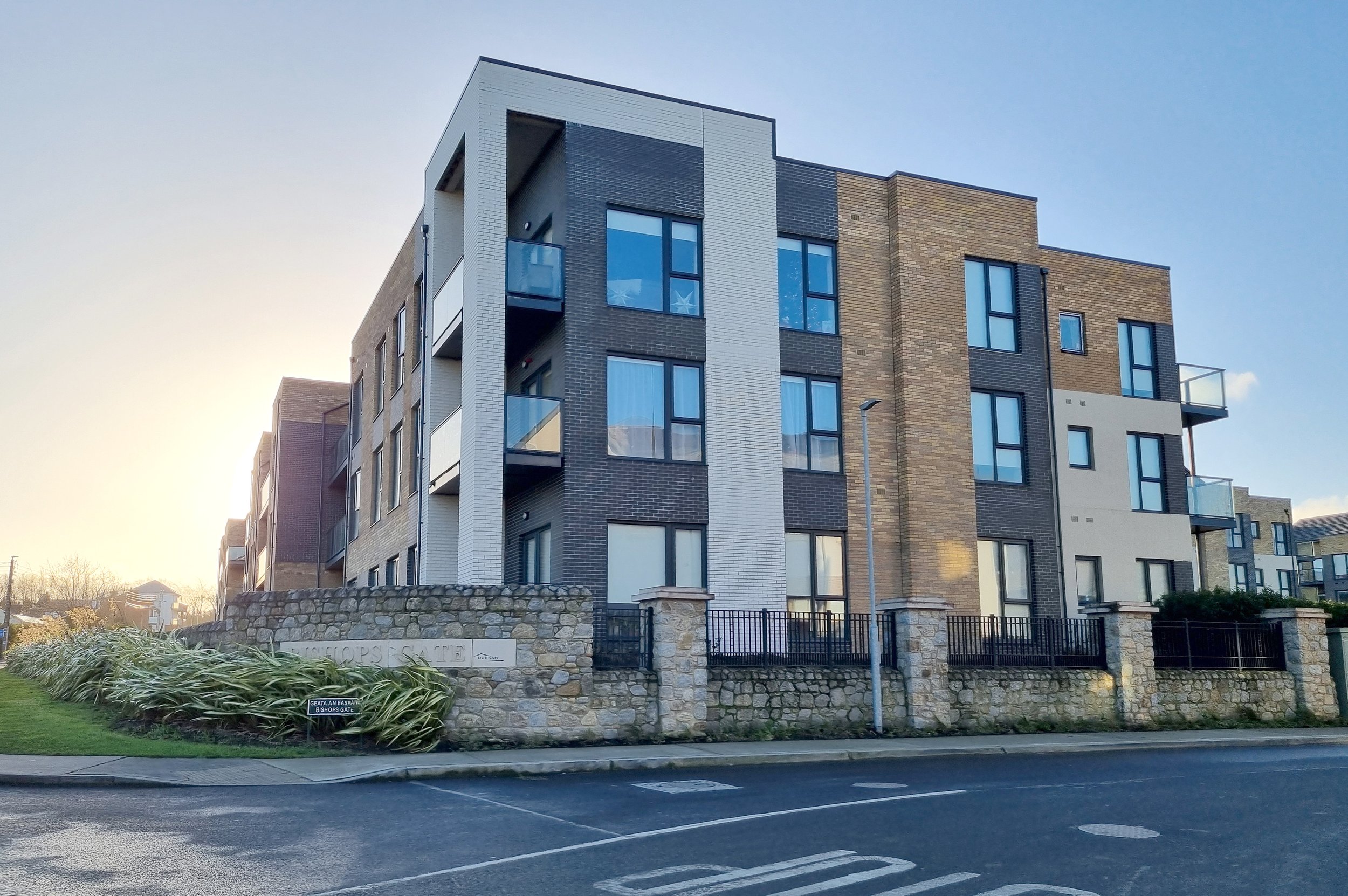 Modern multi-story apartment building with brick and white siding, glass balconies, and a stone wall at the entrance, situated along a paved street.