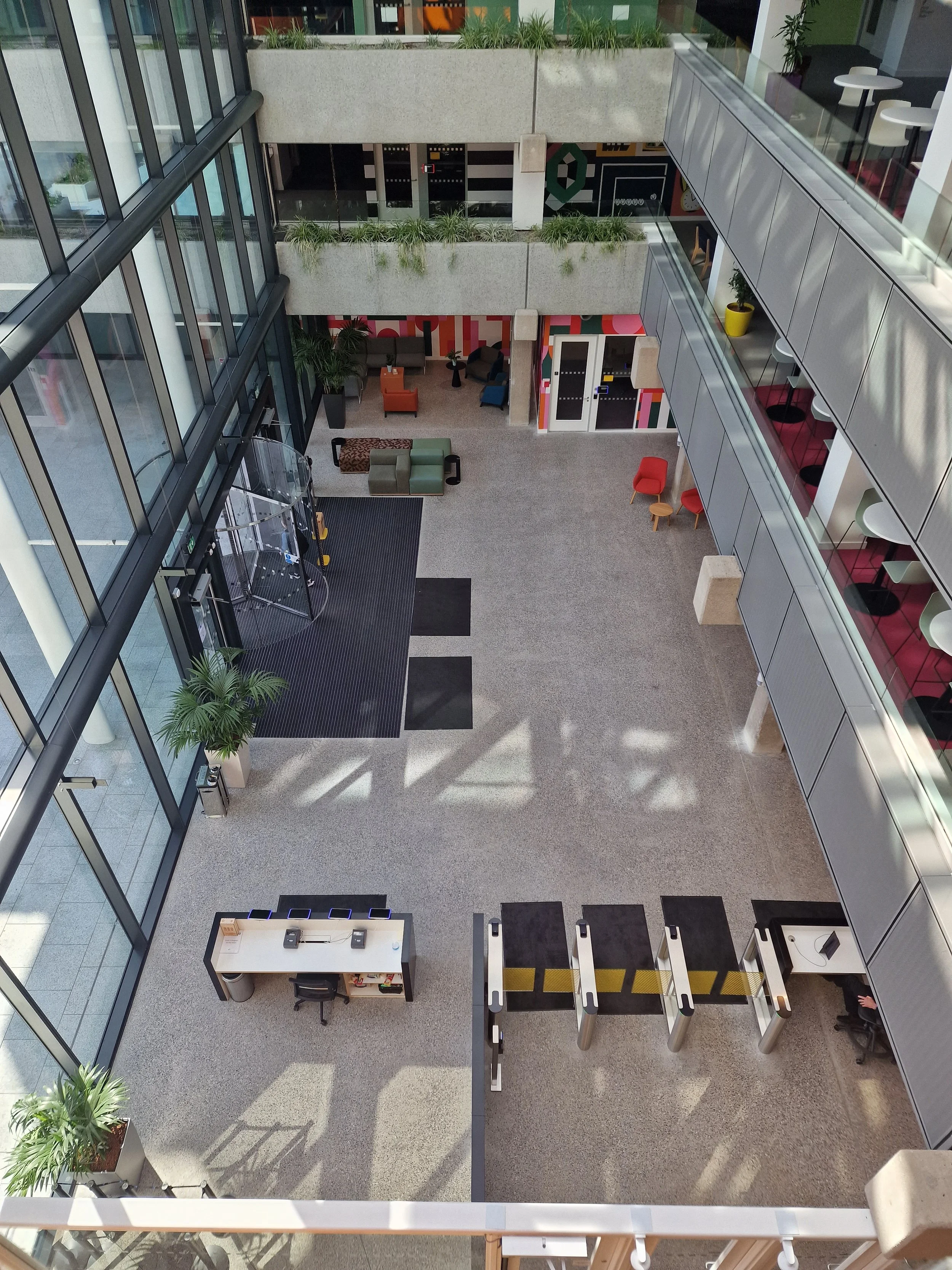 Interior view of a modern building lobby with glass walls, a security checkpoint with turnstiles, a reception desk with phones and computers, colorful seating areas, and indoor plants.