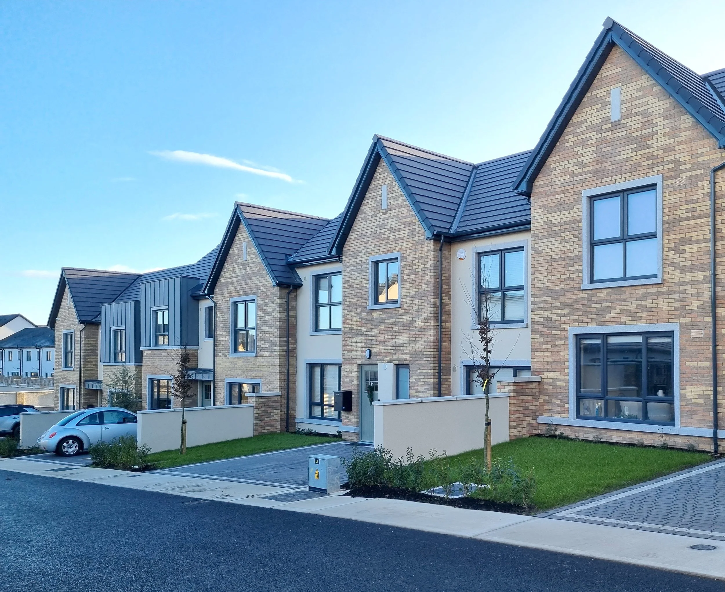 Modern residential townhouses with brick and metal siding, large windows, small front yards, and parked cars on a sunny day.