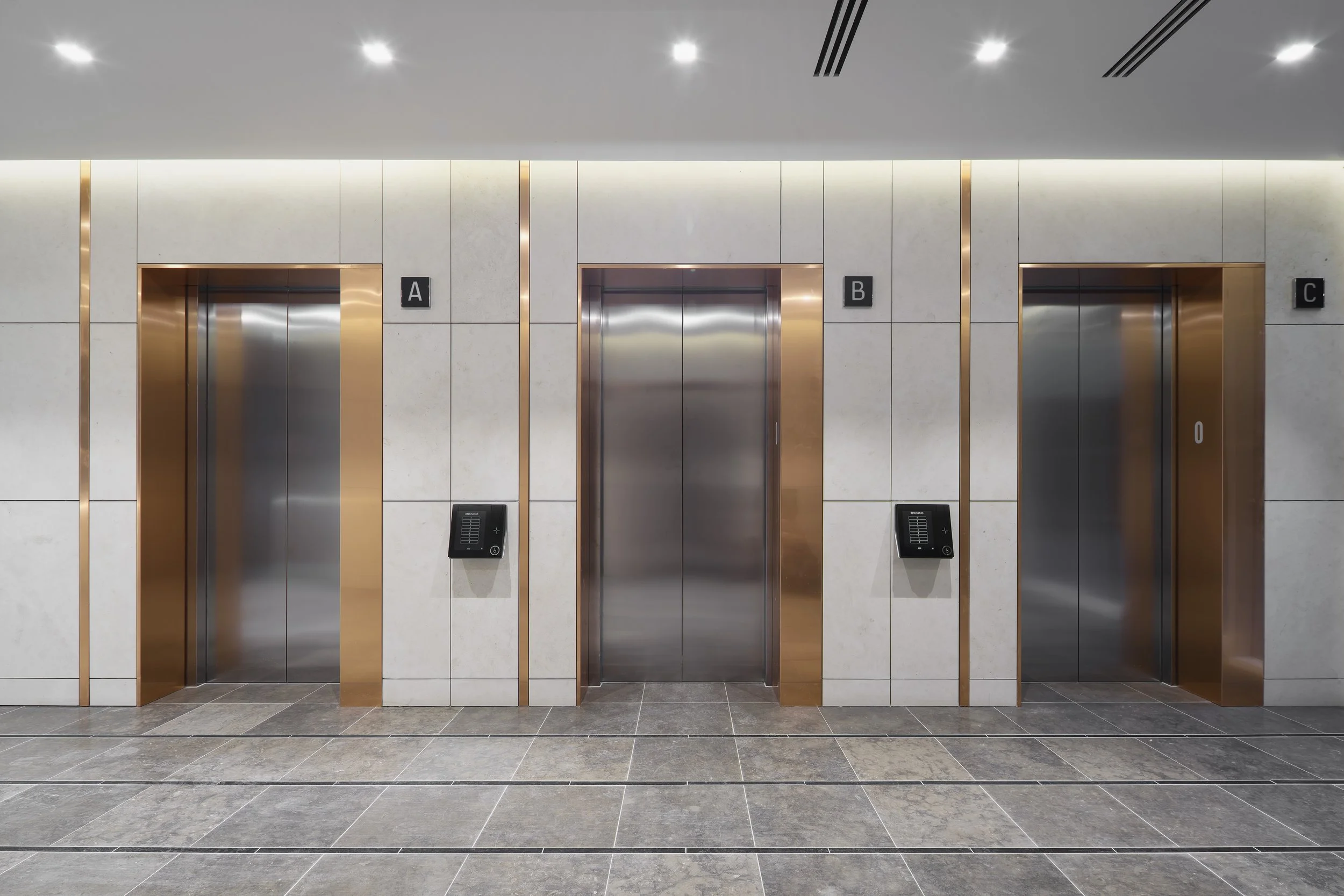 Three elevator doors with metal finishes, labeled A, B, and C, set within a modern building lobby with gray tiled floors and white walls accented with gold trim.