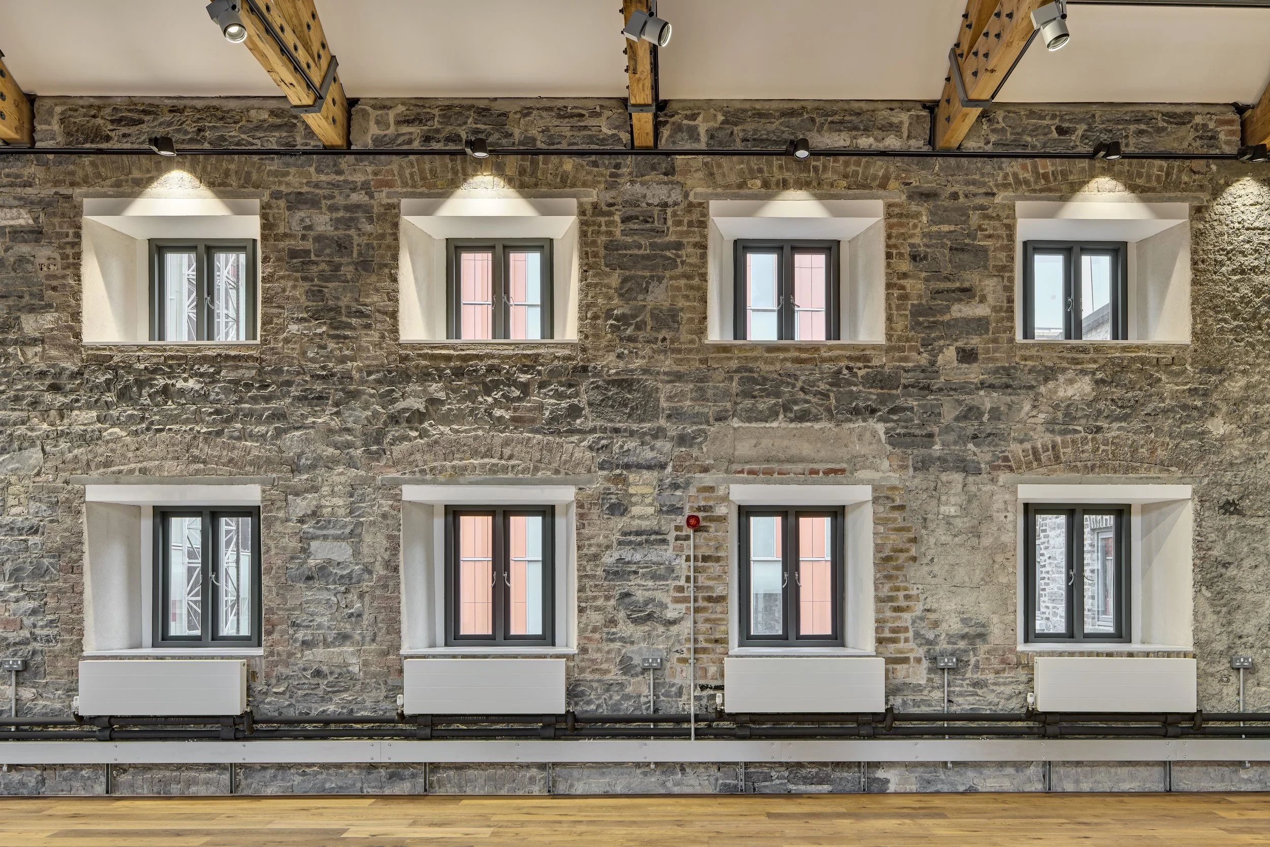 Interior view of a room with six small windows embedded in a rustic brick wall, illuminated by spotlights, with wooden ceiling beams and a wooden floor.
