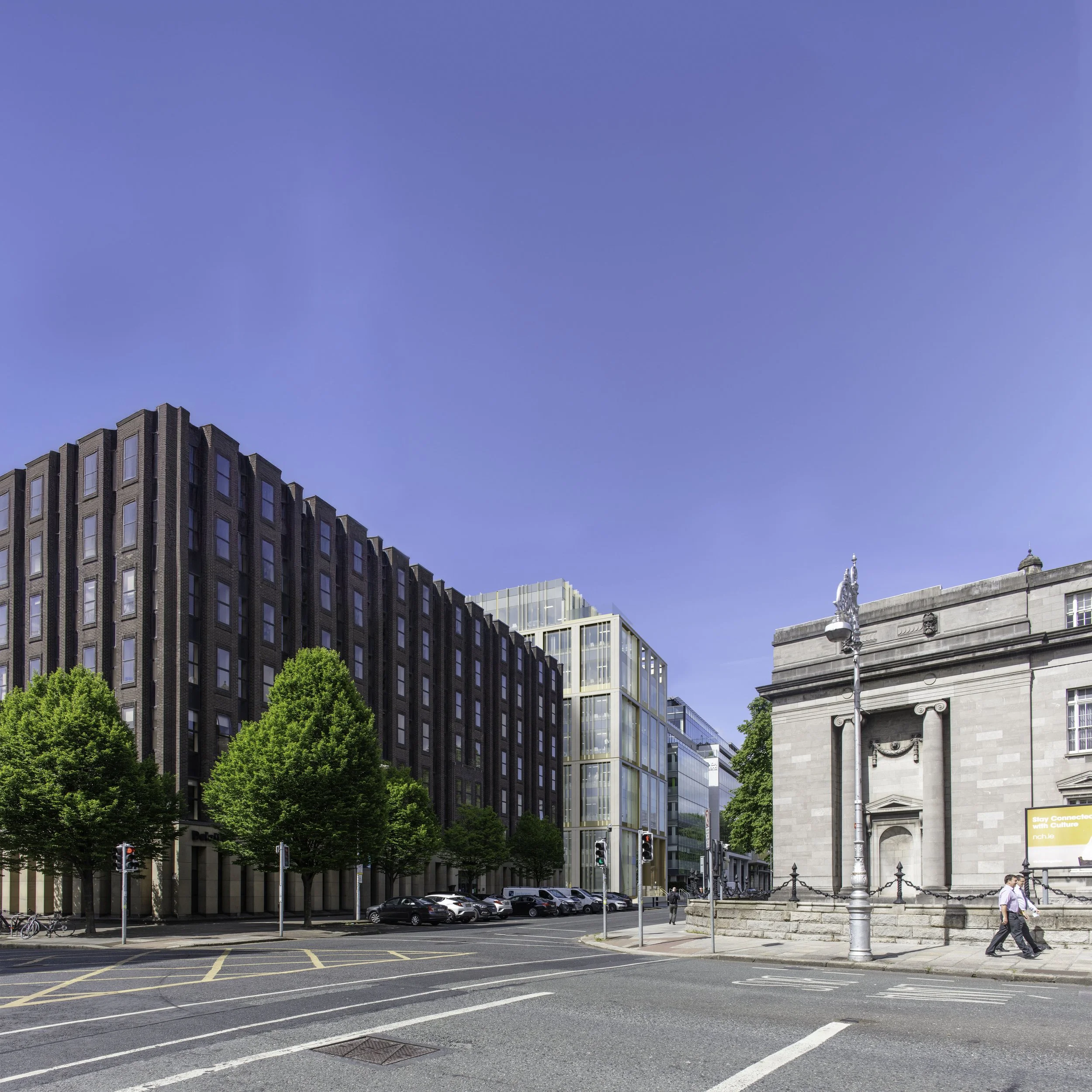 City street scene with modern and historic buildings, cars parked along the curb, trees, traffic lights, and pedestrians walking on the sidewalk on a sunny day.