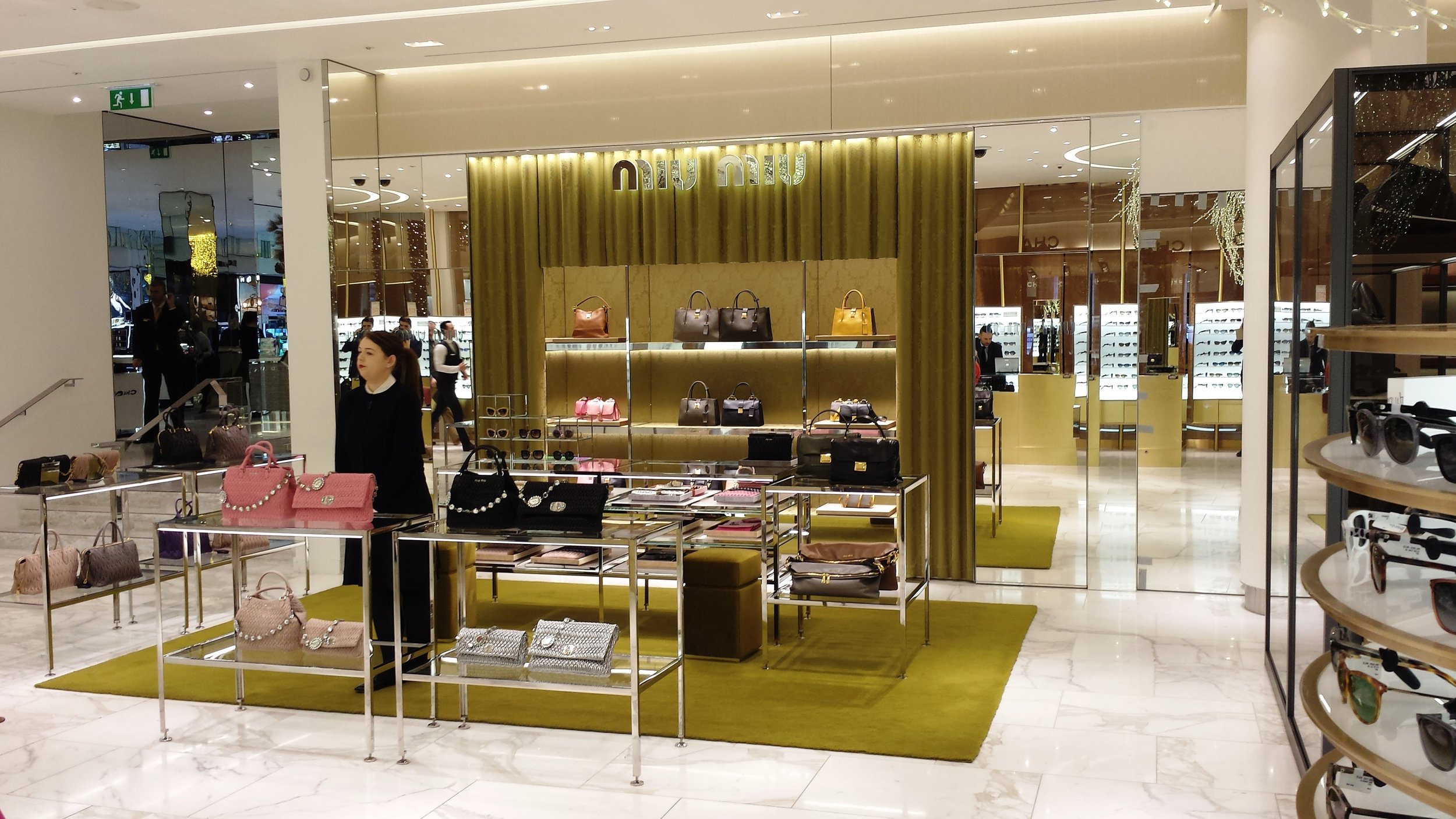 Display of handbags, sunglasses, and accessories in a luxury department store with golden decor, glass shelves, and marble flooring.