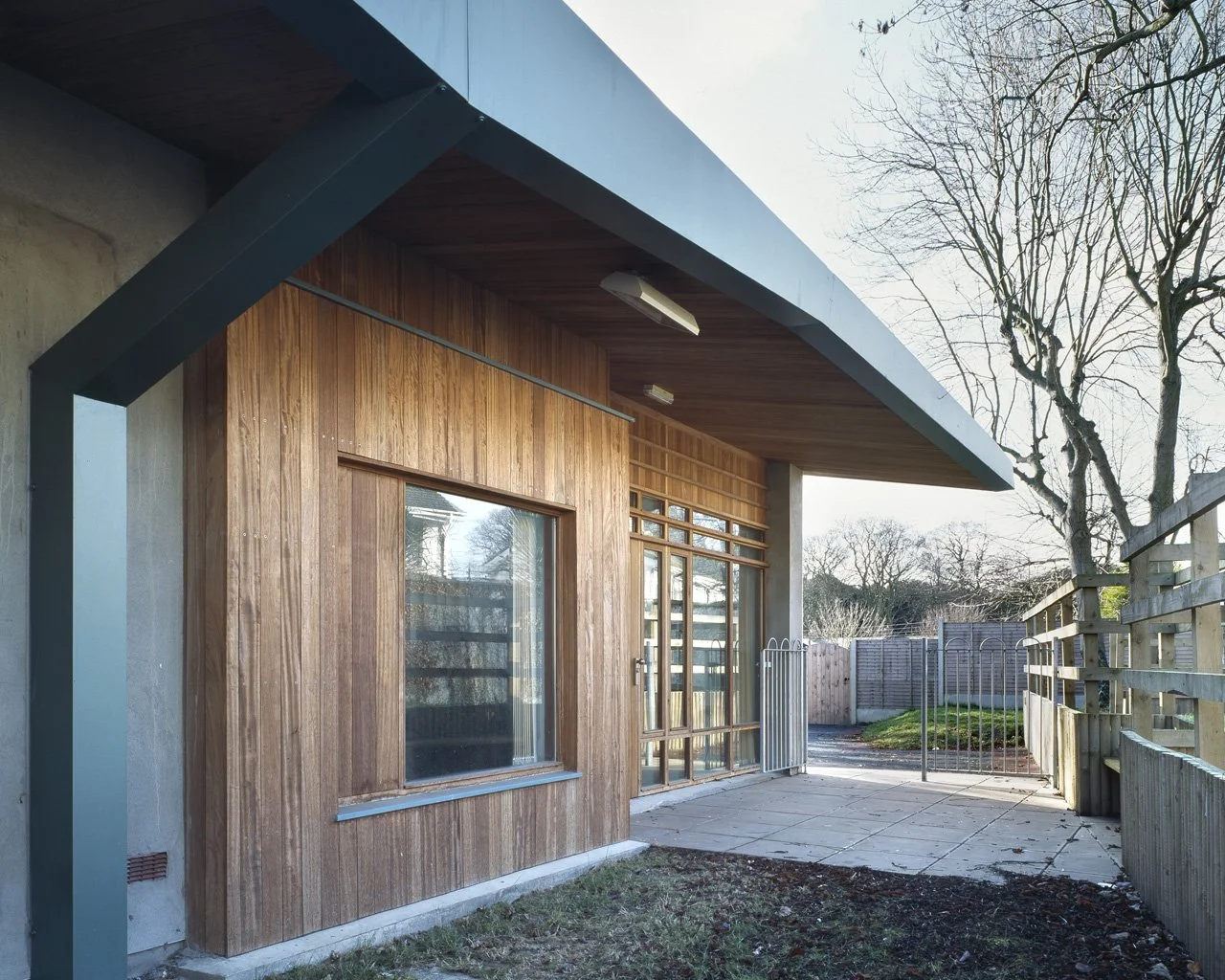 Exterior of a modern building with wooden siding, large windows, a concrete patio, and a fenced backyard with trees.