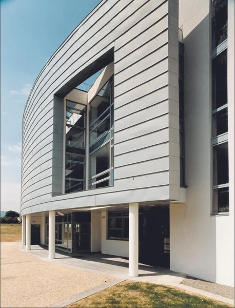Modern building with curved white exterior, large glass windows, and supporting white columns, situated on a grassy area with a partly cloudy sky.