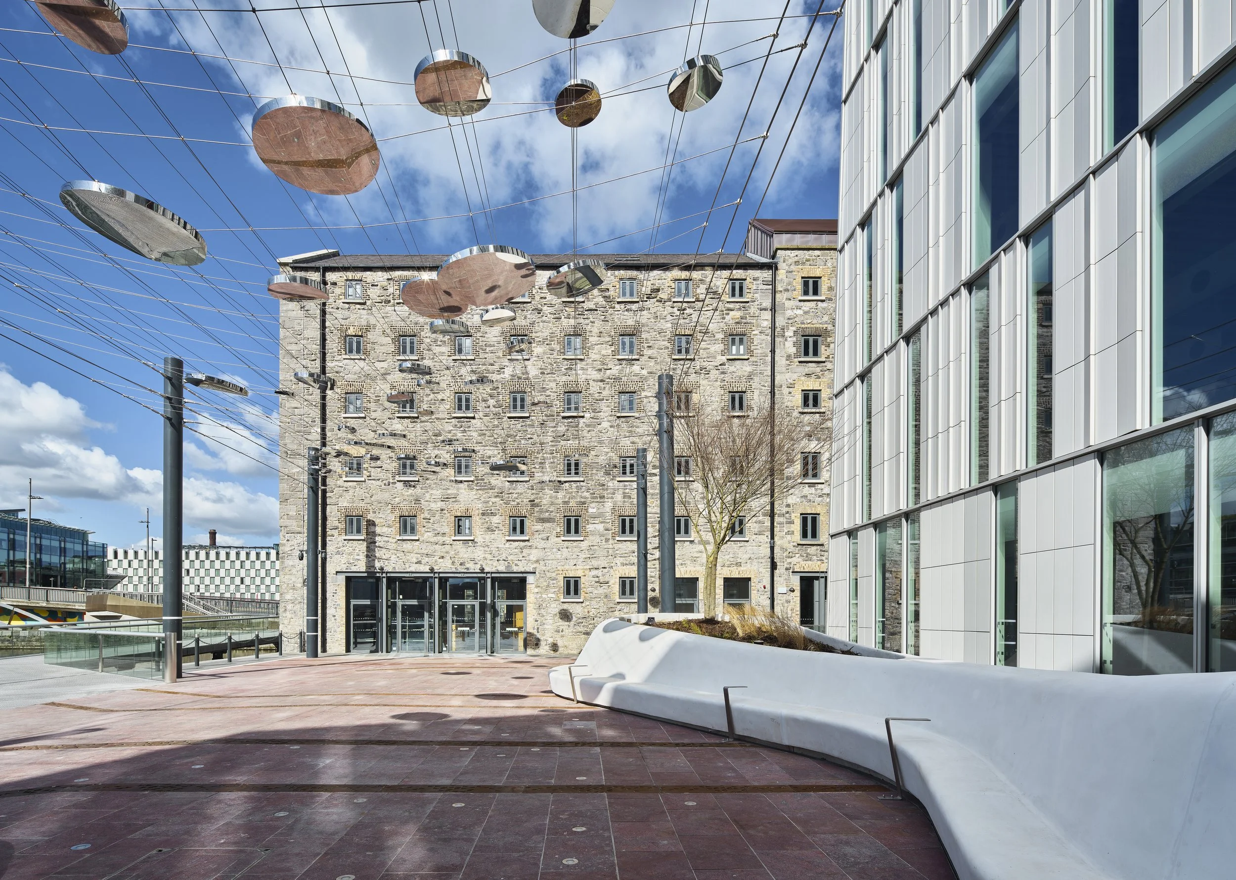 Modern urban architecture with an old stone building, contemporary glass facade, and suspended reflective discs under a blue sky.
