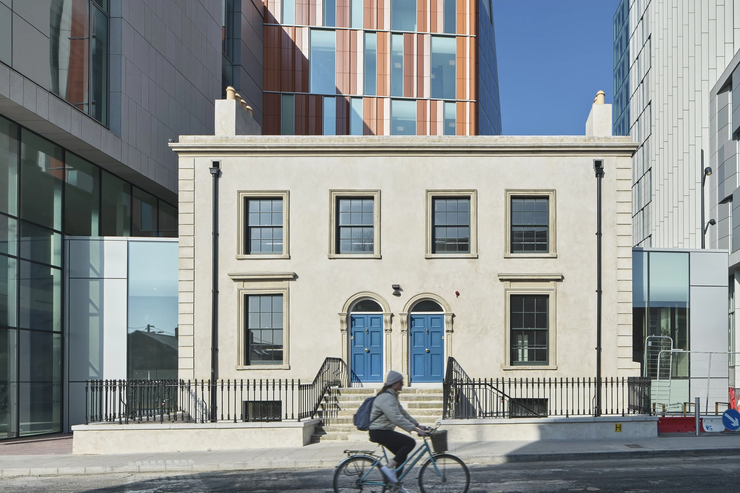 A person riding a bicycle past a historic two-story building with blue doors and multiple windows, surrounded by modern glass and steel buildings in an urban setting.