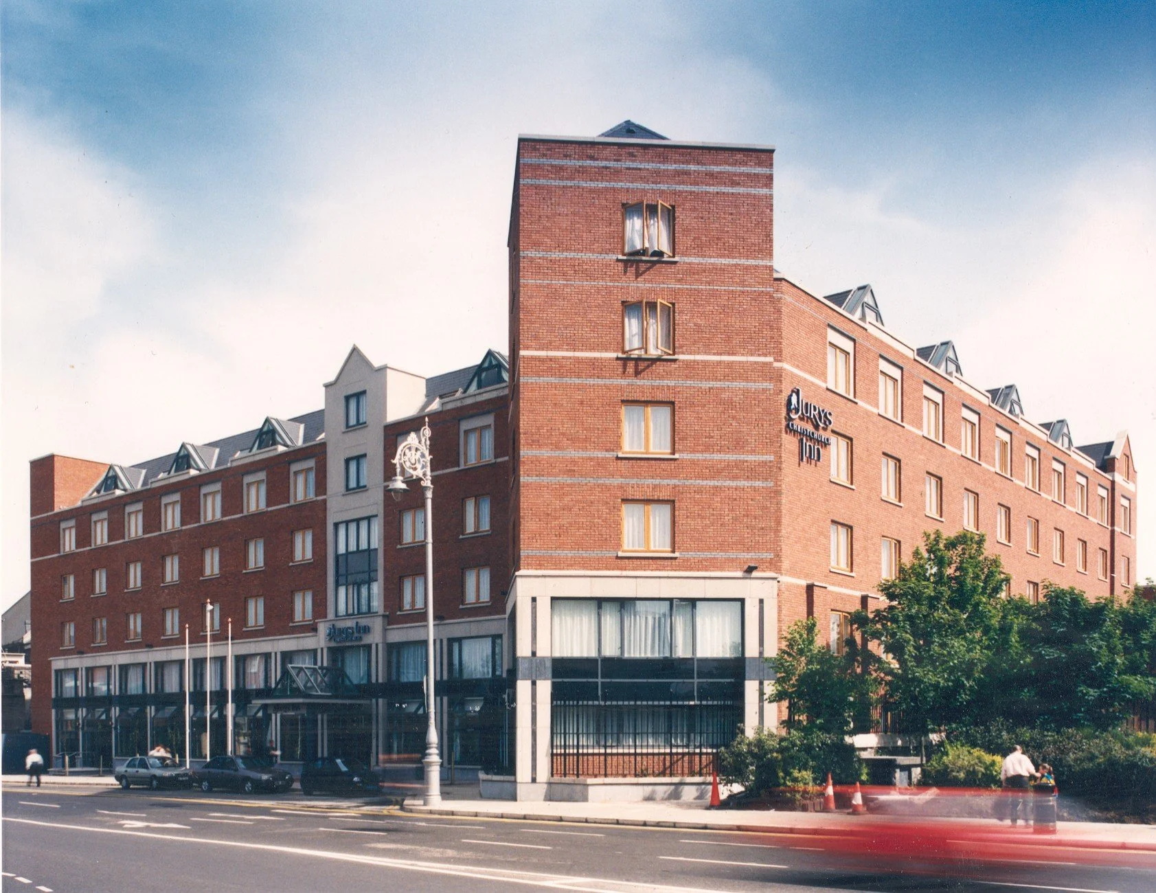 A multi-story brick hotel building with a sign that reads 'Jurys CHICHESTER TOP' on the side. The hotel has large windows, a pitched roof with small dormer windows, and a street view with parked cars, a lamppost, and trees.