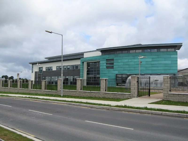 Modern two-story building with teal and gray exterior, surrounded by a black metal and brick fence, located along a paved road under a cloudy sky.