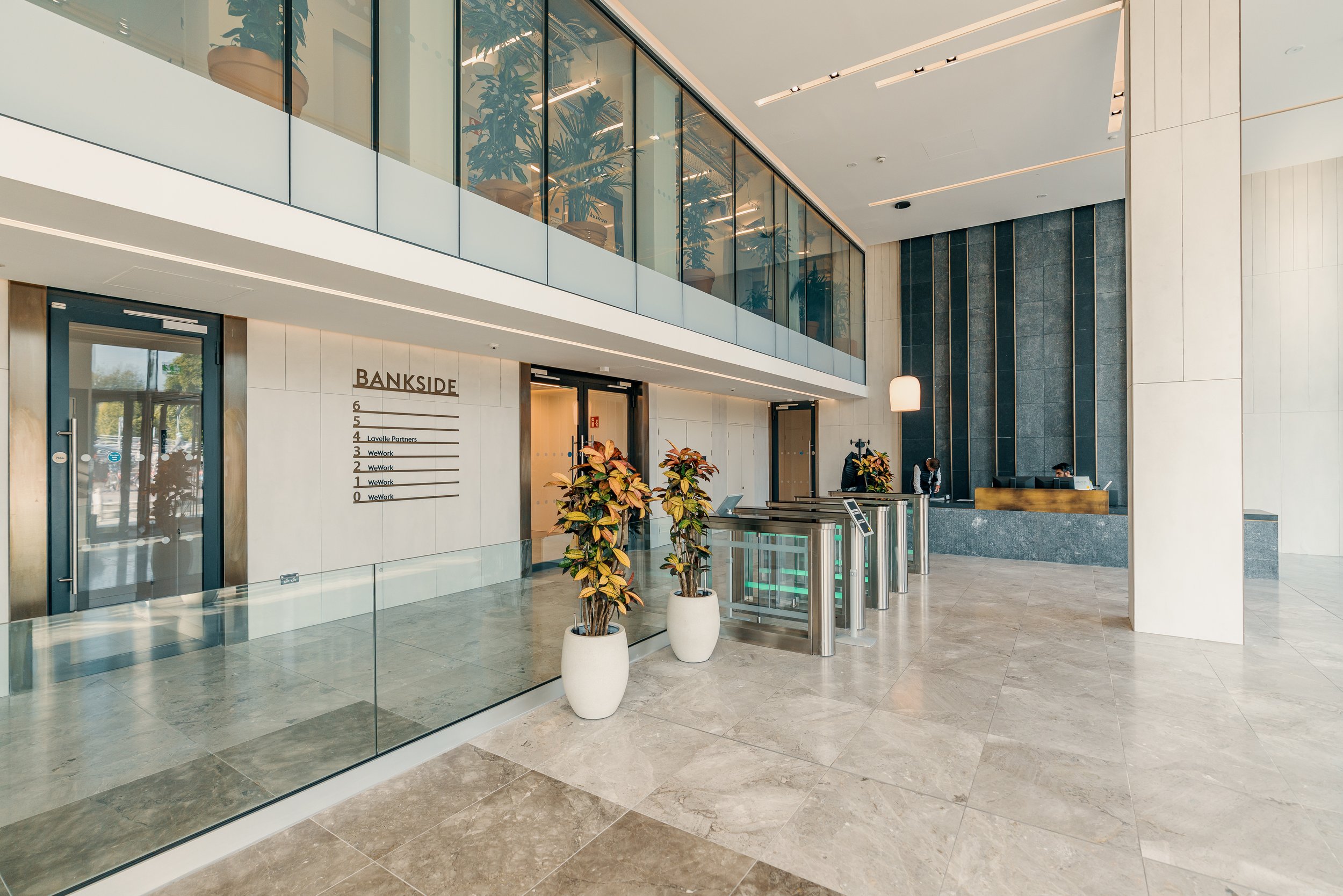 Modern office lobby with a reception desk, potted plants, and a sign displaying office names and floor numbers.