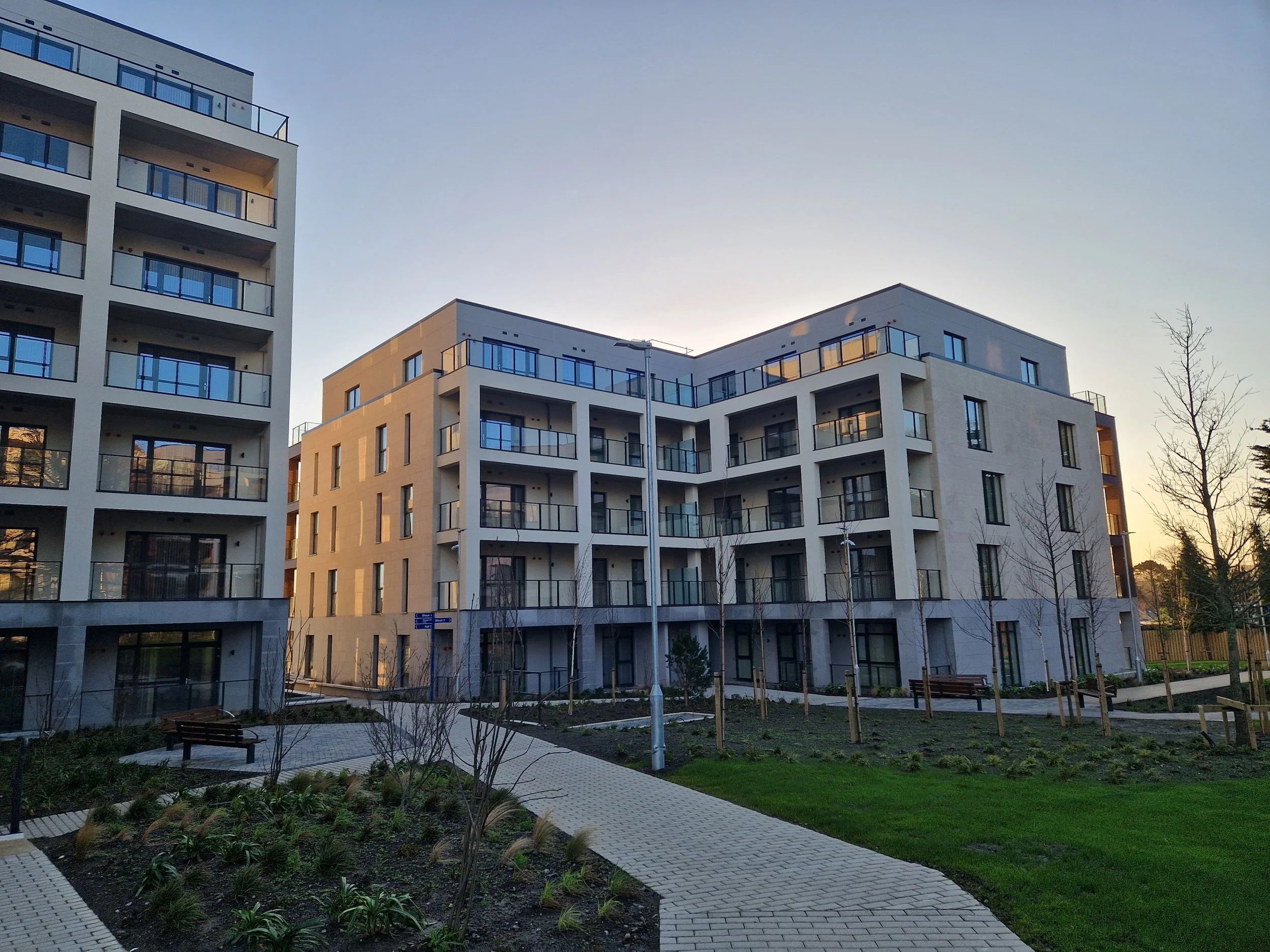 Modern residential apartment buildings with balconies, surrounded by landscaped pathways, benches, and young trees in a landscaped courtyard during sunset.